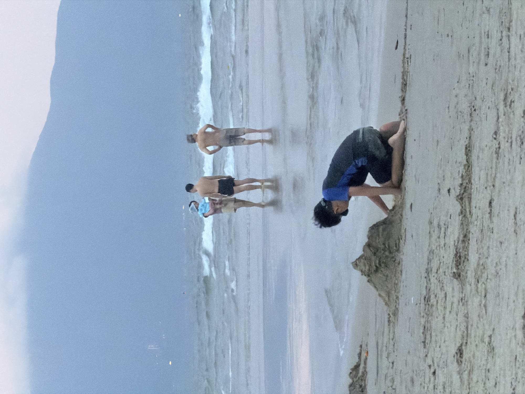 On a beach, presumably in Đà Nẵng, Vietnam, as inferred from the coastal geography and likely tourist activity, four individuals are observed. Three are positioned closer to the water's edge; one appears to be wearing a light-colored shirt, while the other two are shirtless and wearing dark shorts. The fourth individual is situated on the sandy shore closer to the viewer and is bent over, touching the sand with their hands. The sea has visible waves and a low horizon suggests the possibility of an overcast sky or early morning. The beach itself shows a mix of wet sand near the water and drier sand inland.