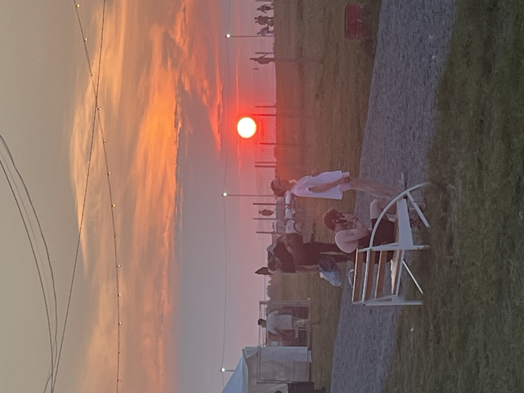 A sunset scene is captured in Surakovo, Russia, with a vibrant orange and pink sky. In the foreground, a person sits on a white and wooden bench, facing away from the camera. To the left, a white tent structure stands. A gravel path extends across the middle ground, with several people walking or standing on it. A tall fence with vertical posts runs through the scene. In the background, more figures are visible, and the sun is a bright red orb near the horizon. String lights are strung across the sky above the scene. The ground is covered in grass and gravel.