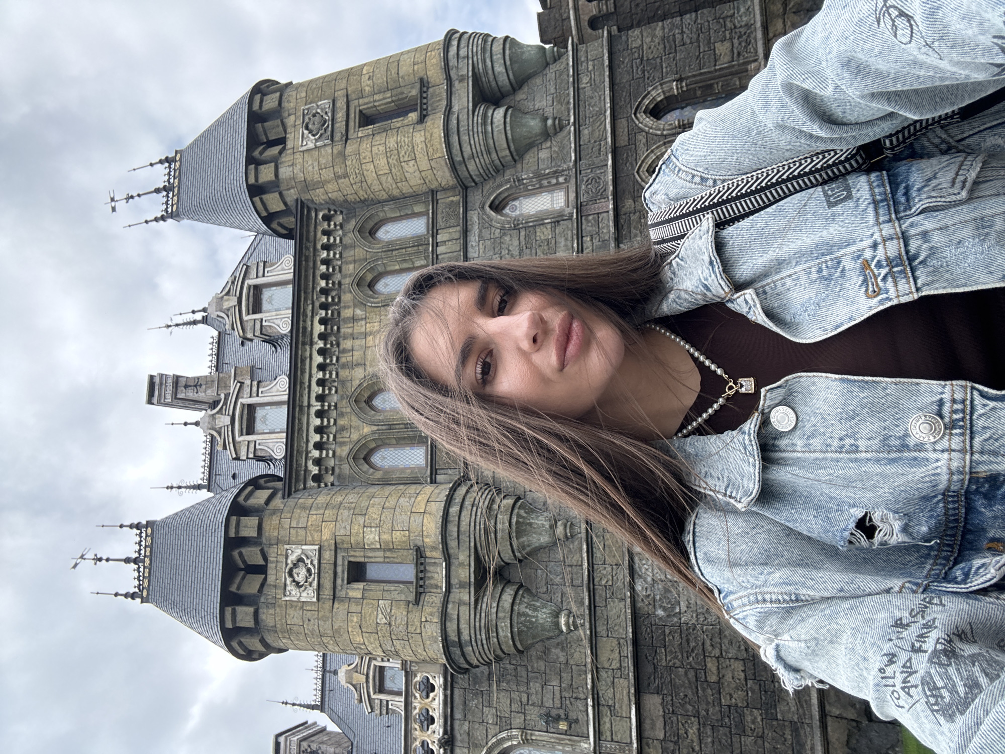 In an outdoor, low-angle selfie, a young woman with long, brown hair stands before a large, gothic-style castle. The woman is positioned in the foreground, facing the camera with a slight smile. She is wearing a distressed denim jacket over a brown top and a pearl necklace. The castle, characterized by its stone construction, pointed roofs, and numerous windows, dominates the background. The sky above is overcast. The castle is identified as the 'Fairytale Castle' in Khryashchevka, Russia. The scene suggests a tourist or leisurely outing at a historical or themed architectural landmark.