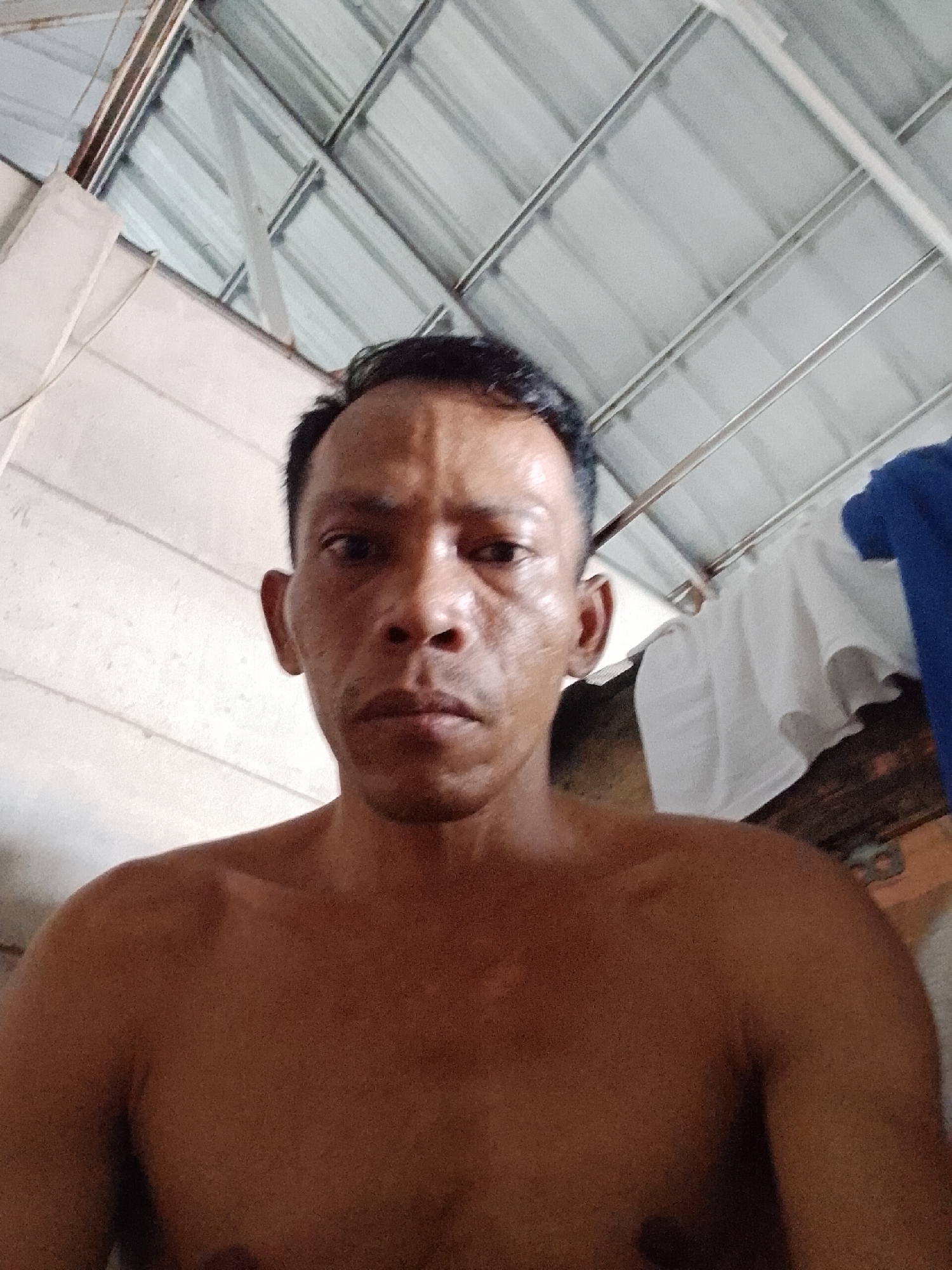 A medium-shot, upward-angled selfie shows a shirtless Asian man with short dark hair and medium-brown skin. He has a serious expression. Behind him and to the left is a white wall. Above him is a ceiling composed of white panels and metal support beams. To the right of the man, partially visible, are folded clothes, one blue and one white. The photo appears to have been taken indoors in Bukit Ketok, Indonesia.
