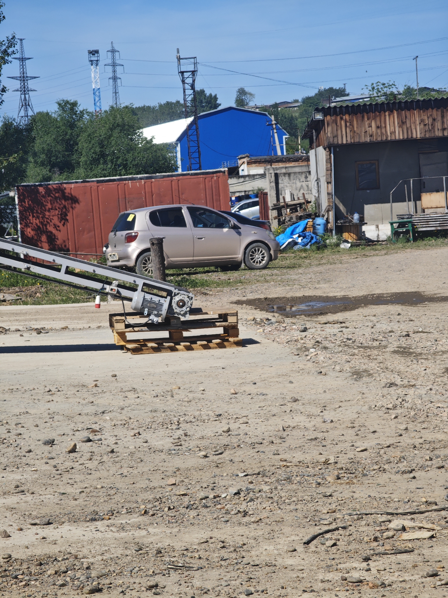 The image depicts an outdoor industrial or storage area on a clear, sunny day. In the foreground, a conveyor belt system is positioned on wooden pallets. Further back, a beige hatchback car is parked next to a large, rust-colored shipping container. Beyond that, a bright blue building with a white roof is visible, along with a weathered wooden structure. The background features a line of trees and several tall electricity pylons. The ground is a mixture of dirt, gravel, and a small puddle. The scene is located in the city of Krasnoyarsk, Russia. There are no people visible in the image.