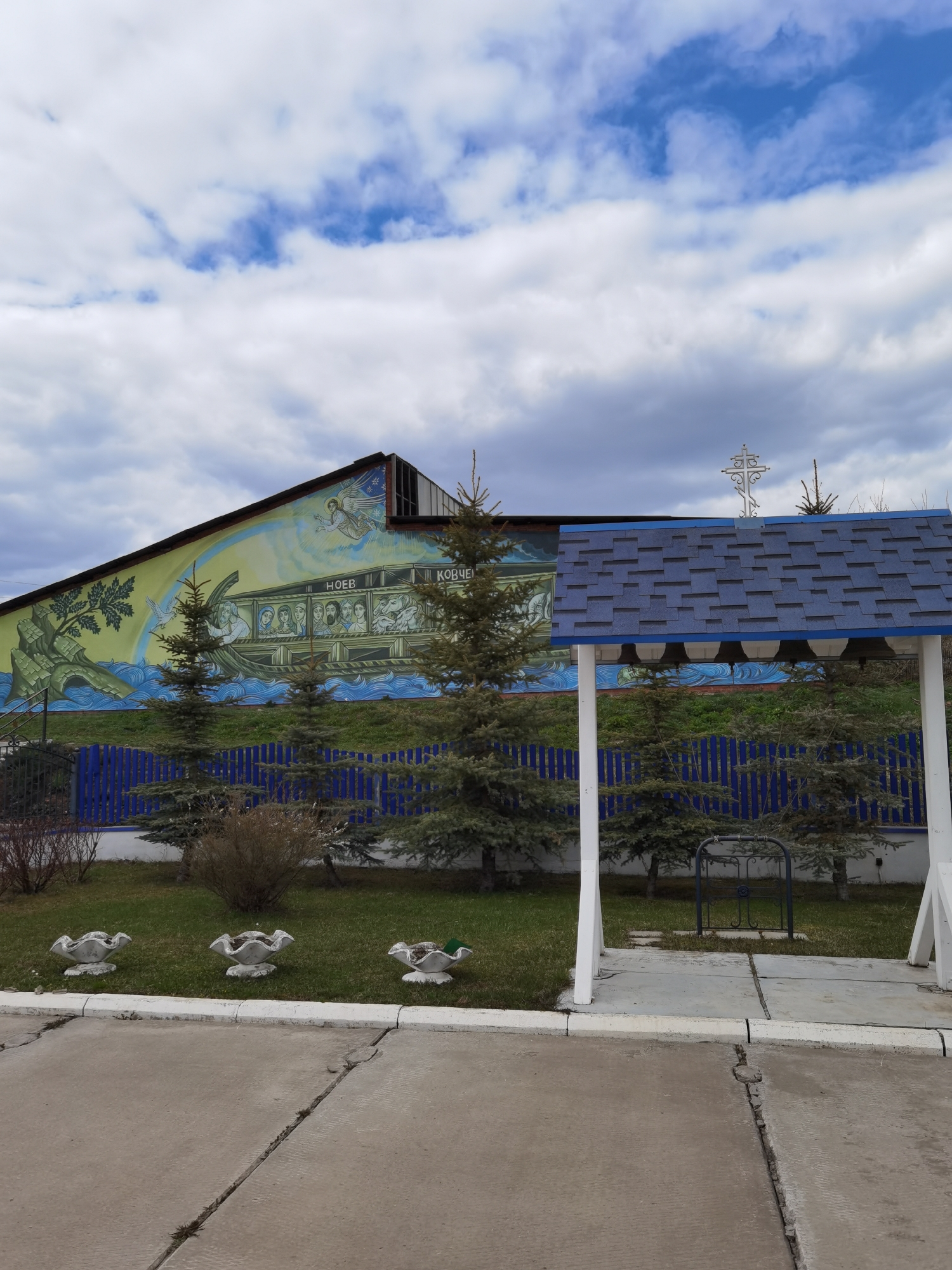 The image shows a building in Кварса, Russia, featuring a mural of Noah's Ark. In front of the building are young evergreen trees and ornamental concrete planters. A small structure with a blue shingled roof and white supports sits in the foreground. A metal cross is visible on top of the building. The sky is overcast with blue patches. The ground is paved.