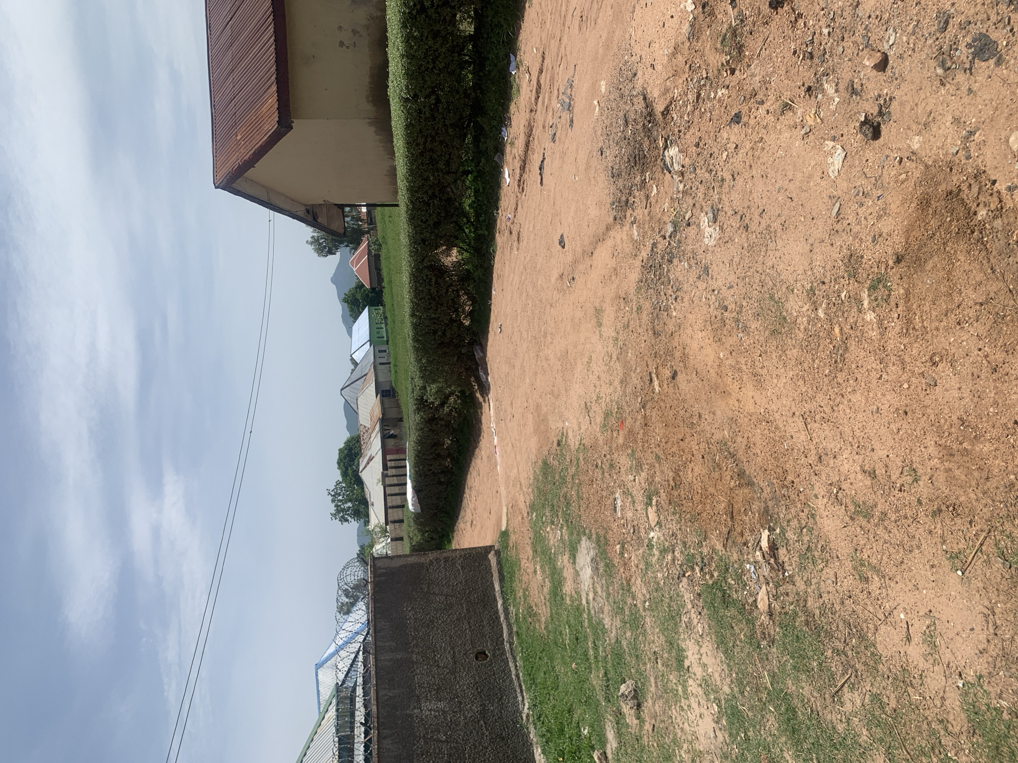 The image depicts a dusty, dirt road leading through a residential area in Jauro Yinu, Nigeria. The road is bordered by a low, neatly trimmed hedge on the right, separating it from a row of simple houses with corrugated metal roofs. On the left, a tall, rough-textured concrete wall topped with barbed wire encloses a grassy area. The sky is partly cloudy, and distant hills are visible. The scene appears to be a snapshot of everyday life in a developing community, characterized by its unpaved roads, modest housing, and natural surroundings.