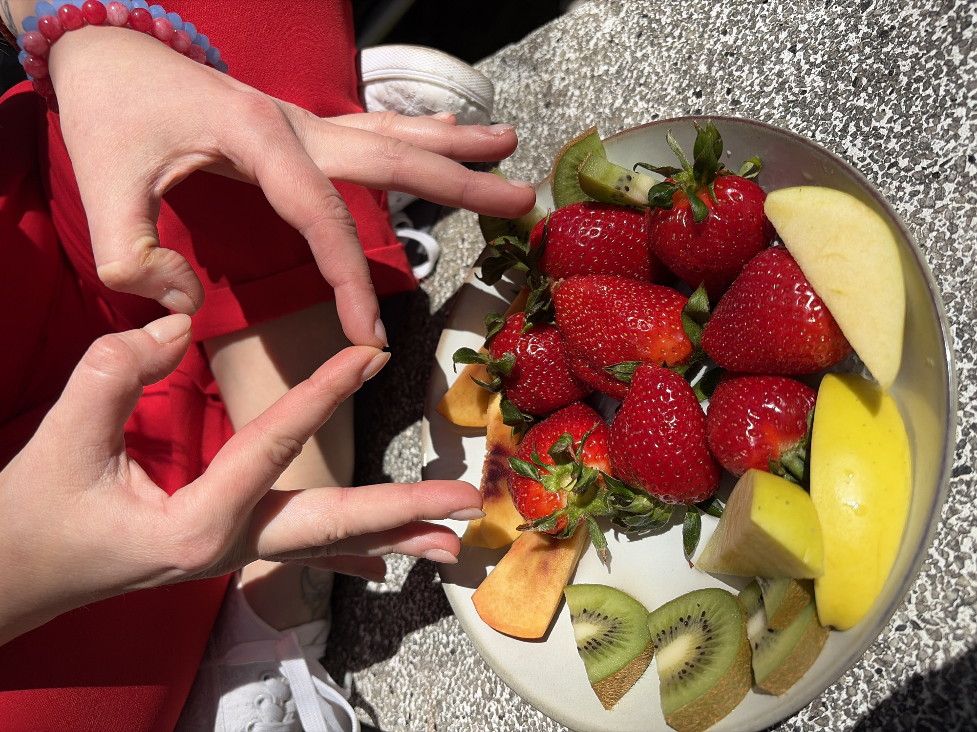 A close-up shot from above shows a person sitting, with a white bowl of fruit on their lap. The person is wearing a red outfit and a blue and red beaded bracelet on their left wrist. Their hands are shaped to form a heart. The bowl contains strawberries, kiwi slices, peach slices, and apple slices. The person is wearing white sneakers. The background is a gray, mottled surface, possibly stone or concrete. The image appears to have been taken outdoors in daylight, but the location is not identifiable as Budva, Montenegro based solely on the visual details.