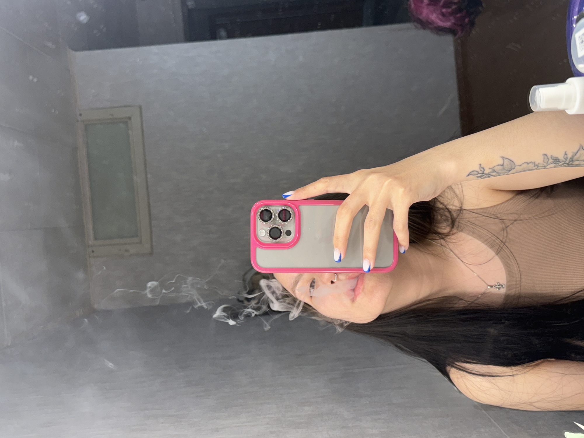 A mirrored selfie depicts a woman with long black hair, wearing a tan top and a silver necklace with a cross pendant. She is holding a pink-cased smartphone with her left hand, and smoke is exhaling from her mouth. The mirror reflects a neutral-toned wall with a recessed panel or frame. A partially visible bottle is positioned at the top right of the frame. The image location cannot be determined to be Фоки, Russia based on the visual content.