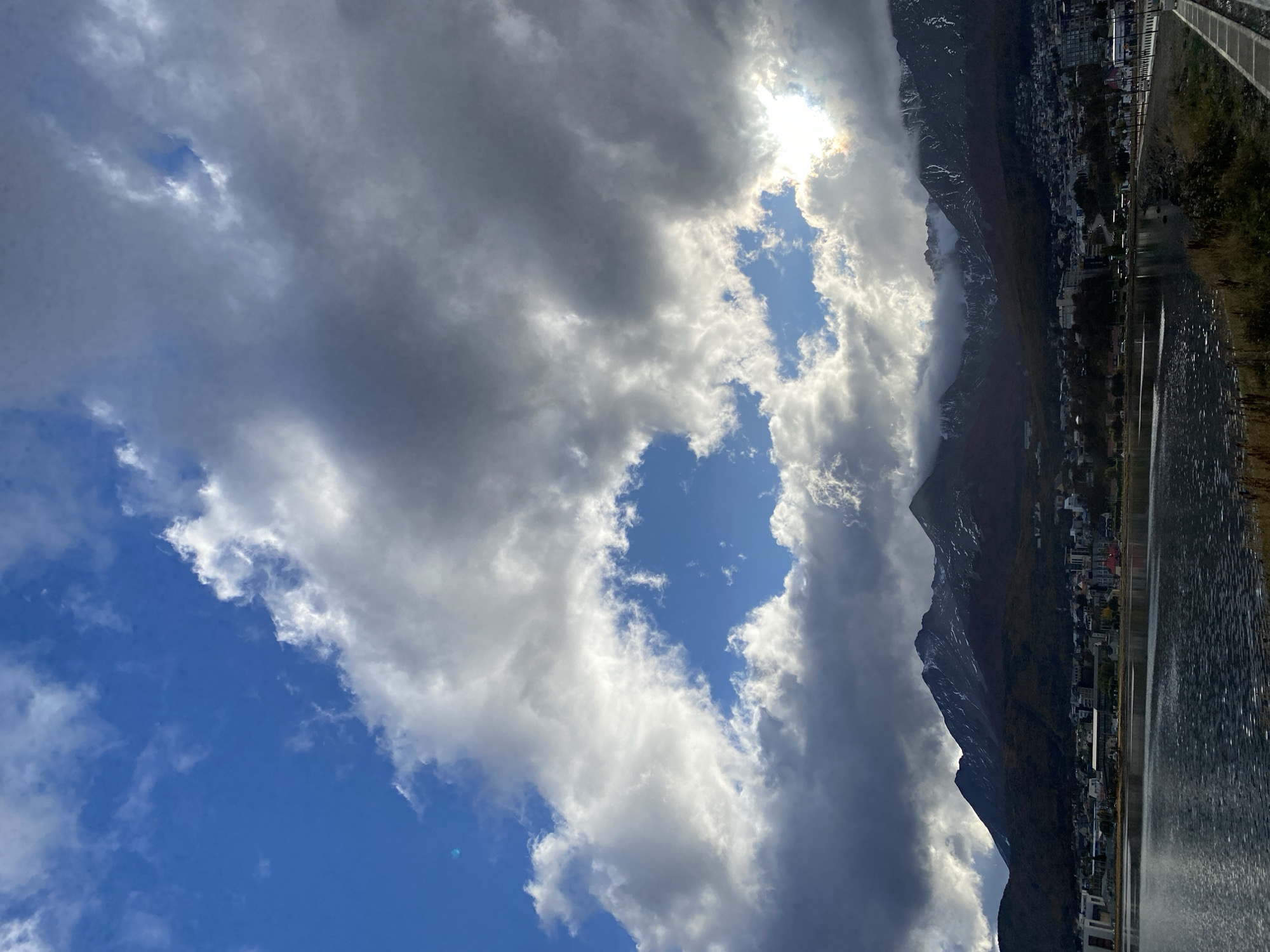 The image shows a view of Ushuaia, Argentina, from a slightly elevated position. The sky is partially covered with clouds, with patches of blue visible. Sunlight streams through some of the cloud formations. A mountain range flanks the right side of the frame, adjacent to a body of water reflecting the landscape. A town or city is nestled between the mountain and the water. A paved pathway runs alongside the water's edge.