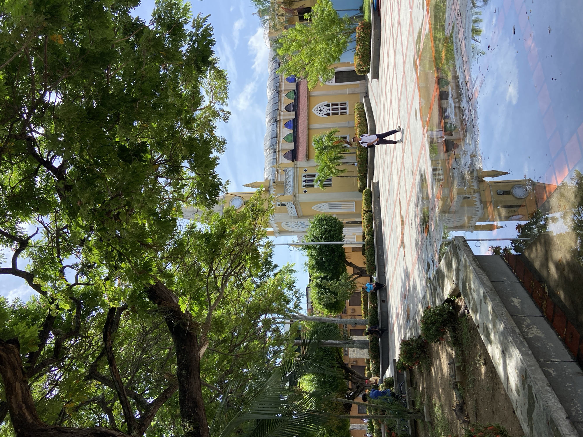 The image captures a wide shot of a town square in San José de Barlovento, Venezuela. A prominent yellow church with a clock tower dominates the background, its reflection visible in a large puddle on the tiled ground. Lush green trees, including palm trees, provide shade throughout the square. A young person, dressed in a white t-shirt and dark pants, walks across the square. Several other individuals are seated on benches around the perimeter of the square, some appearing to be engaged in conversation or simply relaxing. The sky is clear with scattered clouds, suggesting a pleasant day. The paving stones of the square are interspersed with red tiles, creating a decorative pattern. The overall scene depicts a tranquil public space within the town.