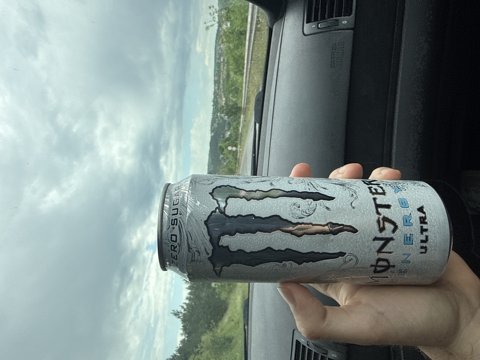A hand holds a can of Monster Ultra Zero Sugar energy drink inside a car. The car's dashboard is visible, and the exterior landscape of trees and sky suggests a road trip. The location cannot be determined as Novi Pazar is a city in both Bulgaria and Serbia.