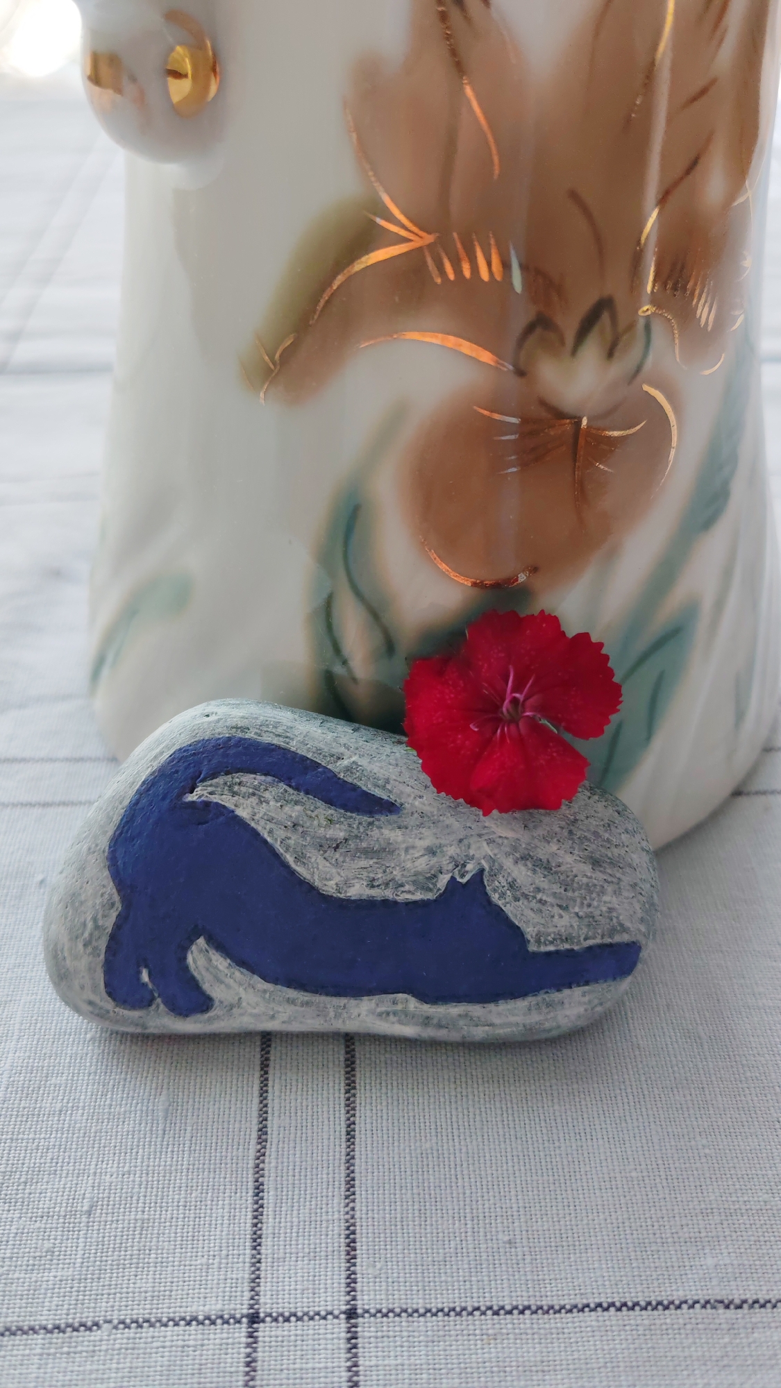 A close-up, eye-level shot shows a painted rock with a blue rabbit design and a small red flower on top of it, placed in front of a white ceramic vase with gold trim and floral painting. The vase and rock are on a white and light blue checkered tablecloth. The scene is lit by natural light, creating soft shadows and highlighting the textures of the objects. The background is blurred, drawing attention to the foreground details.