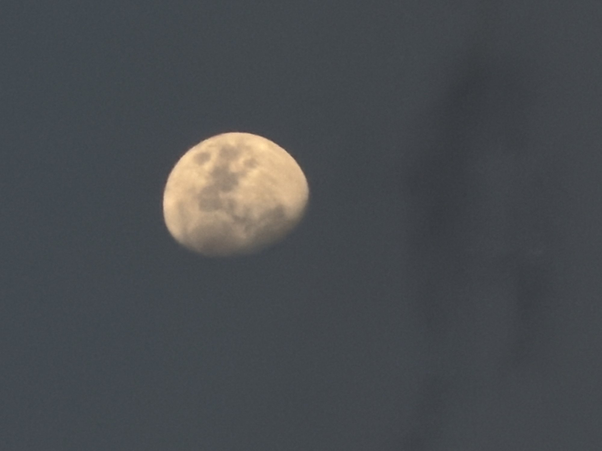 This media file is a close-up shot of the moon against a dark, overcast sky. The moon is mostly illuminated, showing craters and surface details. The sky is a deep gray, suggesting it might be dusk, dawn, or a cloudy day. There are no people or other objects visible in the frame. The lighting is dim, and the overall mood is somewhat subdued due to the lack of bright light and the overcast conditions. No text is visible in the image. The image appears to be a still photograph.
