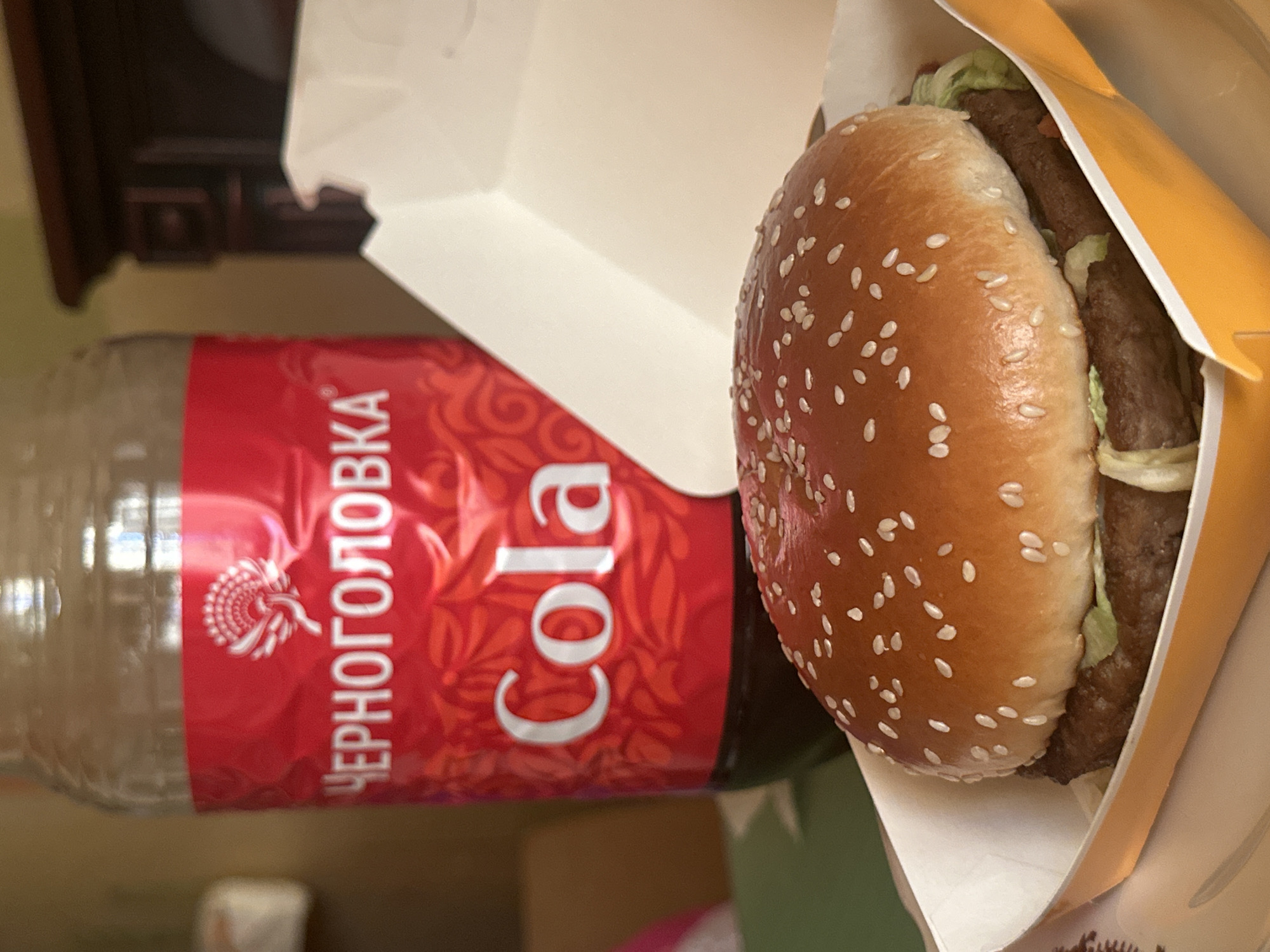 A photograph shows a hamburger in a takeout box and a bottle of Chernogolovka Cola. The cola bottle has a red label with the text "ЧЕРНОГОЛОВКА" and "Cola" in white letters. The hamburger bun is covered in sesame seeds and sits in a partly open yellow and white cardboard box. The environment appears to be indoors, on a surface with a blurred background that includes furniture. While it suggests a food setting, Moscow, Russia, can only be determined through the brand of the cola, which is a Russian brand.
