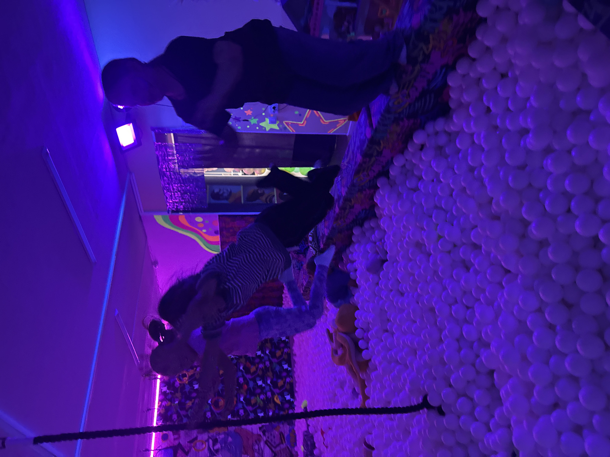 The image depicts a dimly lit indoor scene with vibrant purple and blue lighting, suggesting a playful or entertainment-focused environment. Two individuals, one adult and one child, are present. The adult is partially obscured, bent over, and appears to be interacting with the child. The child, dressed in a striped shirt and patterned pants, is positioned in a ball pit filled with numerous white balls, actively engaged in playing. The background features colorful, possibly psychedelic, decor, including a patterned fabric with flowers and stars. The overall atmosphere seems energetic and fun, likely an arcade, indoor playground, or a similar recreational space. The time of day and weather are not discernible due to the indoor setting and artificial lighting. No text is visible in the image.