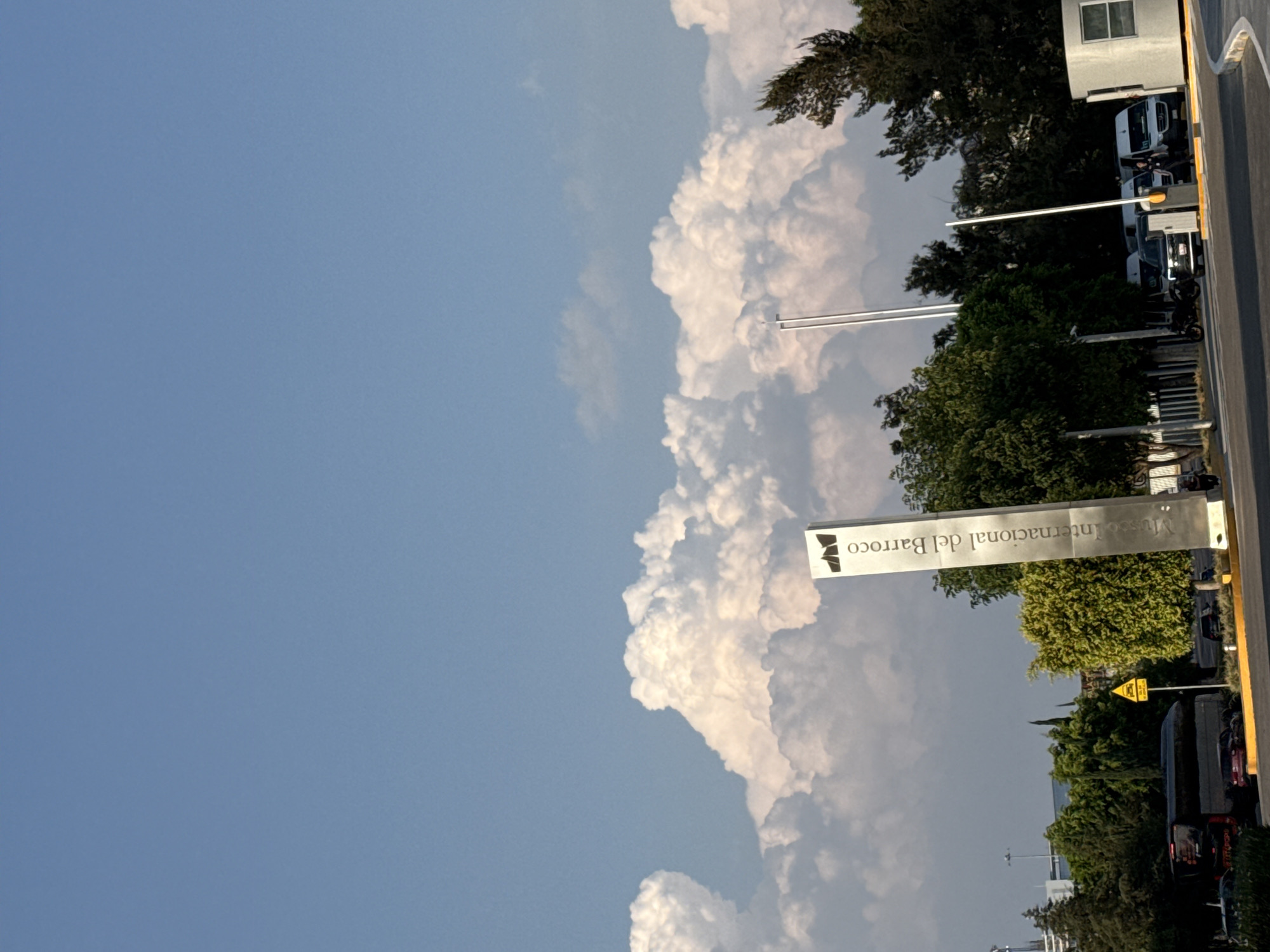 The image captures an outdoor scene in San Bernardino Tlaxcalancingo, Mexico, showcasing the Museo Internacional del Barroco. The museum's signage is prominently visible amidst lush greenery and parked vehicles. The sky is a mix of clear blue and billowing white clouds, creating a dynamic backdrop. The presence of trees, a building, and infrastructure suggests an urban or suburban setting. The composition of the image provides a glimpse into the cultural and environmental landscape of the location.