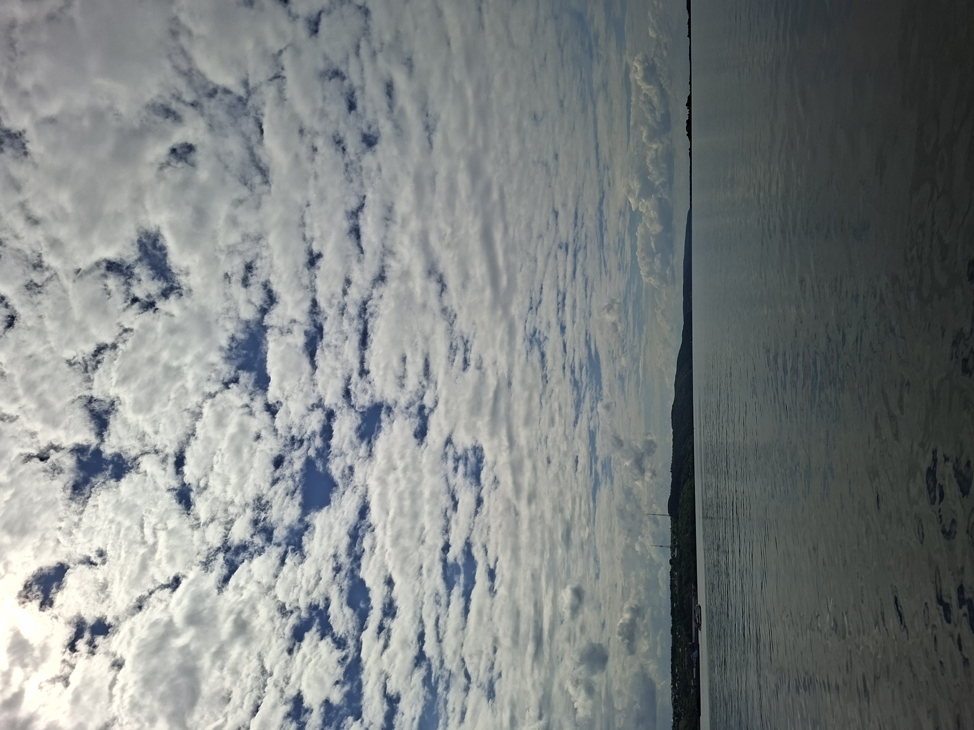 This is an image of a wide, calm body of water, likely a river or lake, under a cloudy sky. The horizon is visible, with a treed, hilly landscape along the distant shore. On the left side of the shore, there are some buildings and a small boat. Two tall, thin towers are visible on the hills in the distance. The water surface shows gentle ripples. The sky is filled with a mix of white and gray clouds, with patches of blue sky visible. The overall impression is of a peaceful, natural scene.
This scene is likely the Volga River in Kazan, Russia. The Volga River is a major waterway that flows through Kazan. The hilly landscape and the presence of buildings and towers along the shore are consistent with the geography of Kazan.