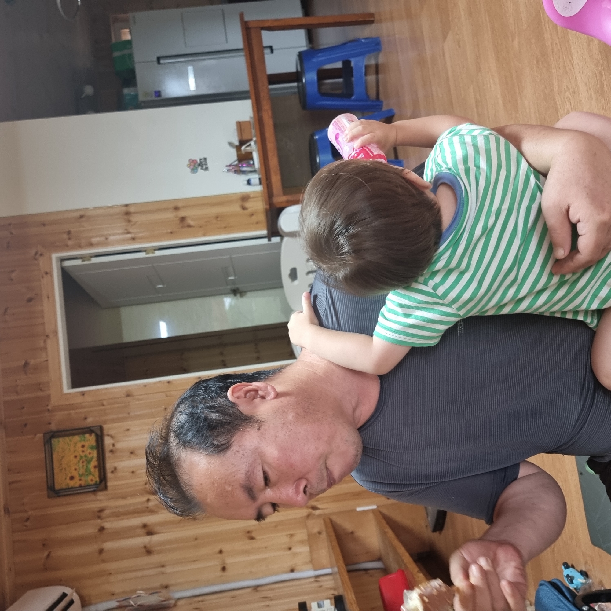 A man and a young child are in a room with wooden walls. The man is seated and holding something edible in his left hand. The child is being held by the man and is drinking from a pink bottle with their right hand. The child is wearing a green and white striped shirt. In the background, there is a wooden table with blue stools underneath, a refrigerator, and a door. The room appears to be a domestic setting.
