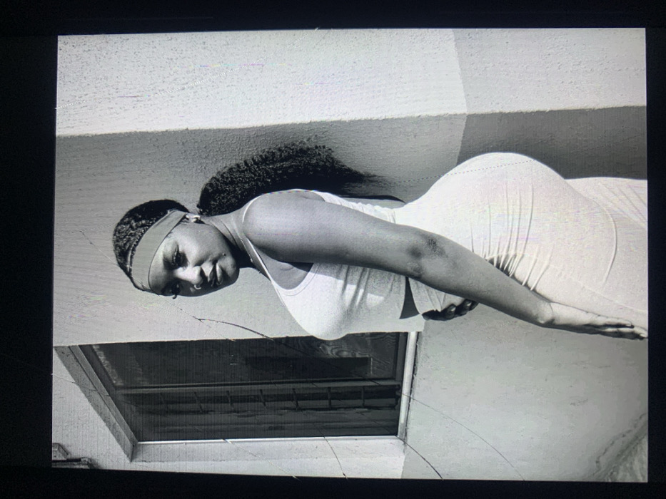 A black and white photograph, captured from a digital screen, shows a single female subject posing outdoors in the city of Jos, Nigeria. The woman, of apparent African descent, is depicted in a three-quarter profile, turned slightly to her left, with her head angled towards the viewer. She wears a light-colored, fitted two-piece ensemble consisting of a sleeveless crop top with spaghetti straps and a matching ruched midi-length skirt. Her dark, braided hair is pulled back into a high ponytail, secured by a light-colored headband. She exhibits a nose ring on her left nostril and an earring on her visible left ear. Her left hand rests on her abdomen, while her right shoulder leans against a light-colored, textured building wall. The background to her left features a window with dark panes and a light frame set into a similar wall. To her right, a plain, light-colored wall shows a distinct vertical shadow, indicating strong directional lighting. The overall scene suggests an exterior urban or residential setting.