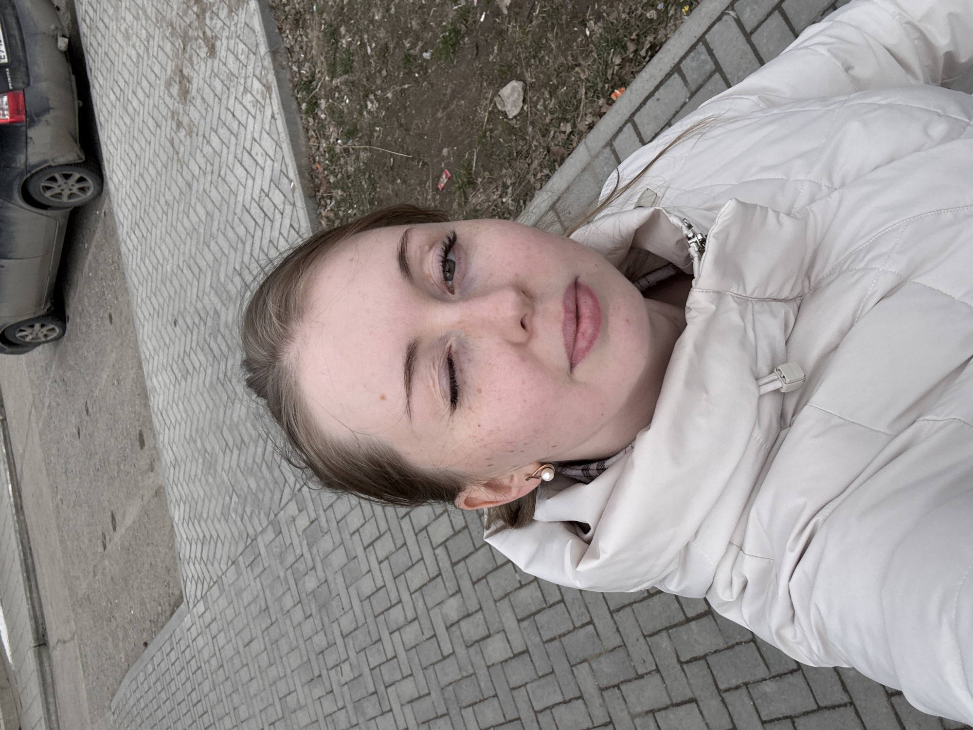 A young woman poses for a selfie outdoors in an urban environment. She is wearing a white puffer jacket and pearl earrings. Her left eye is closed in a wink. She is standing on a paved sidewalk constructed of grey bricks. To the left of the sidewalk is a grey road and a dirty black car is parked. Beyond the sidewalk to the right is a patch of dirt with some green vegetation.