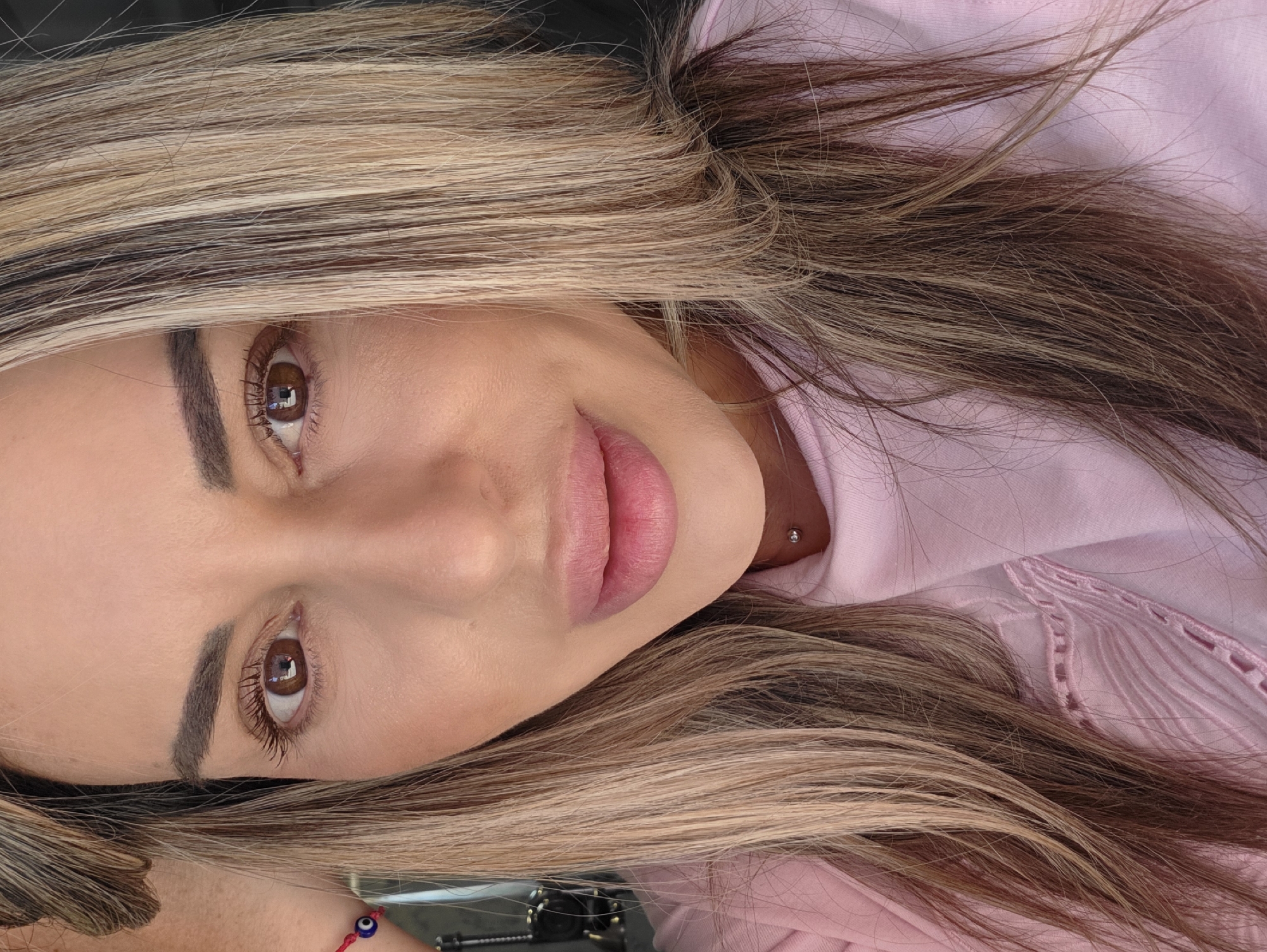 A close-up, head-and-shoulders portrait of a woman with blonde and brown highlights in her hair. She has brown eyes, dark eyebrows, and is wearing pink lipstick. She has a silver stud piercing on her neck. She is wearing a pink shirt. The background is dark and out of focus. No specific location in Carpi, Italy is discernible.