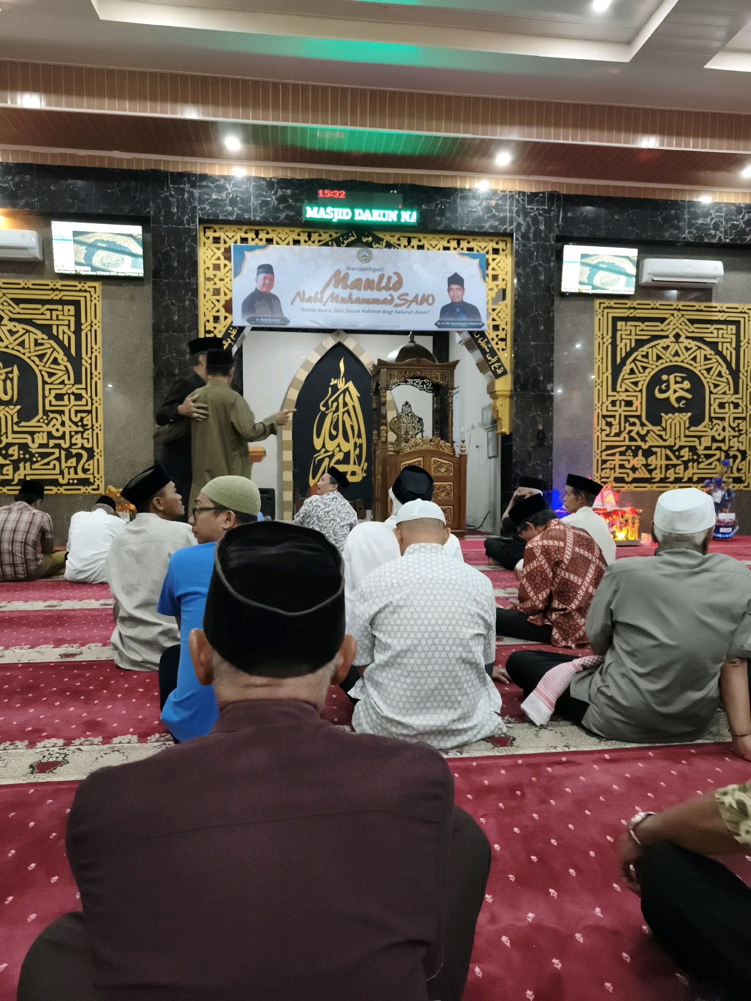 The image depicts an indoor gathering, likely a religious service, taking place within a mosque in Makassar, Indonesia. A large group of men, all wearing caps or skullcaps, are seated on a red carpet. The mosque's interior features traditional Islamic geometric patterns on gold-colored panels adorning the walls. Above the seated attendees, a digital display shows "15:32" and "MASJID DARUN NA'IM," indicating the time and the mosque's name. A banner is visible in the background, featuring portraits of two men and text that appears to be related to a religious commemoration, possibly "Maulid Nabi Muhammad SAW." Several individuals are standing and interacting near a podium or pulpit area. The overall atmosphere suggests a formal religious event.