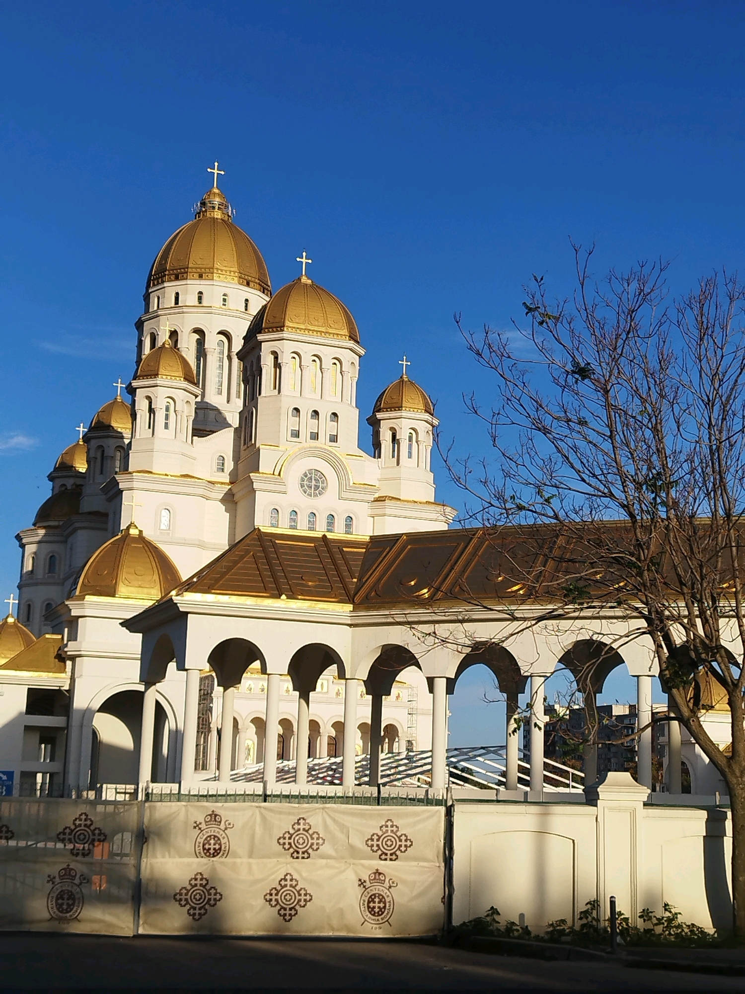 The image shows the exterior of the Romanian Orthodox Cathedral in Bucharest, Romania, under a clear blue sky. The cathedral features multiple golden domes and white architectural elements. In the foreground, a portion of a white fence or barrier with decorative patterns is visible, along with a bare tree branch extending into the frame from the right. The lower part of the cathedral features an arcade with white columns and arches. The overall scene suggests a bright, sunny day.