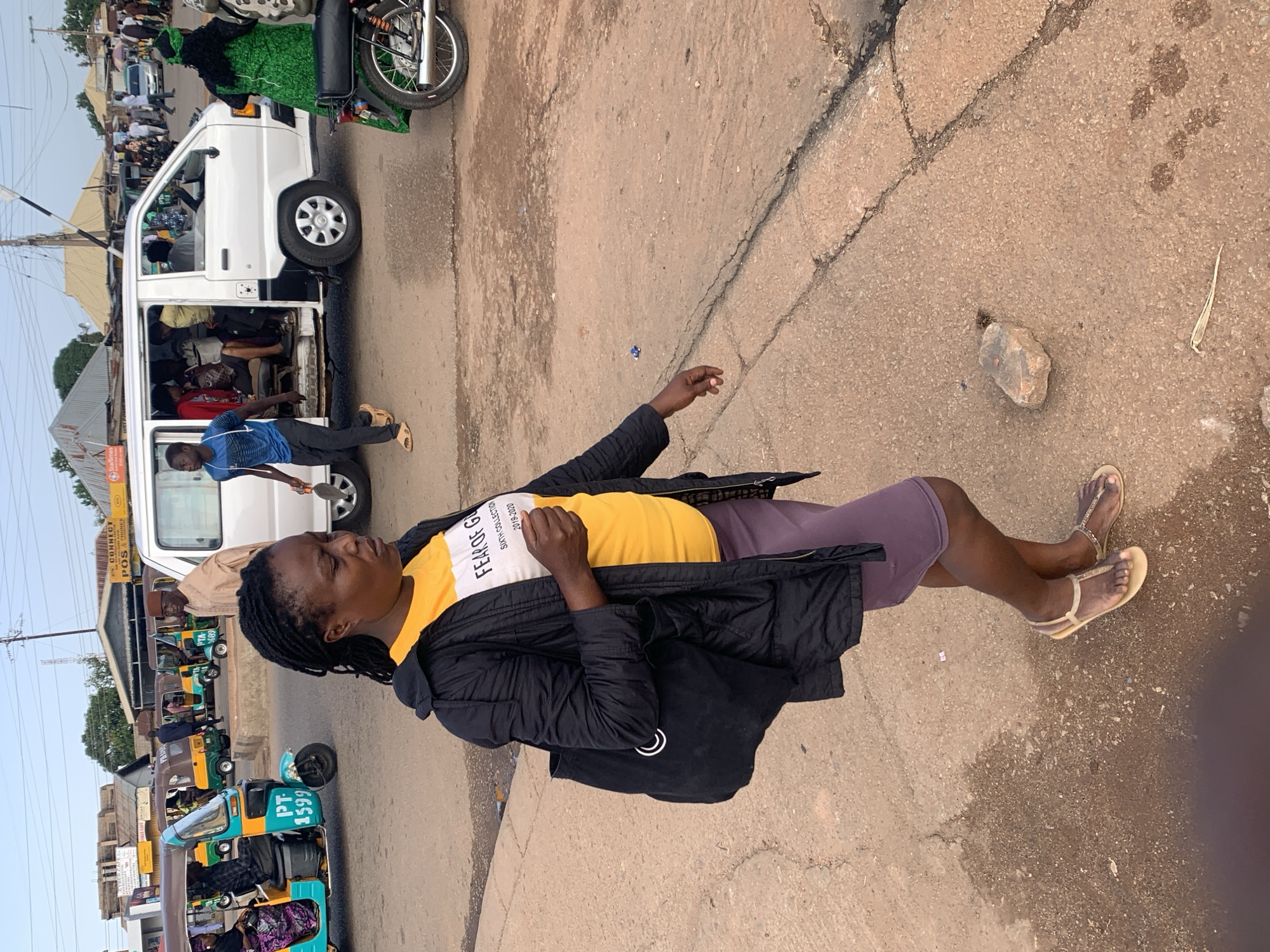 The image depicts a street scene in Gura Top, Nigeria, under an overcast sky. In the foreground, a Black woman with braided hair, wearing a black puffer jacket over a yellow t-shirt with white text reading "FEAR OF GOD" and "SIXTH COLLECTION 2019-2020", and a purple skirt, walks on a paved road, carrying a black tote bag. She is wearing gold flat sandals. Behind her, several auto-rickshaws, known locally as "Keke NAPEP", are parked and in motion. One turquoise and yellow Keke NAPEP has "PT 1599" written on it. A white van is also present, filled with passengers. Other people are visible in the background, along with buildings and market stalls. The ground is a mixture of asphalt and compacted earth.