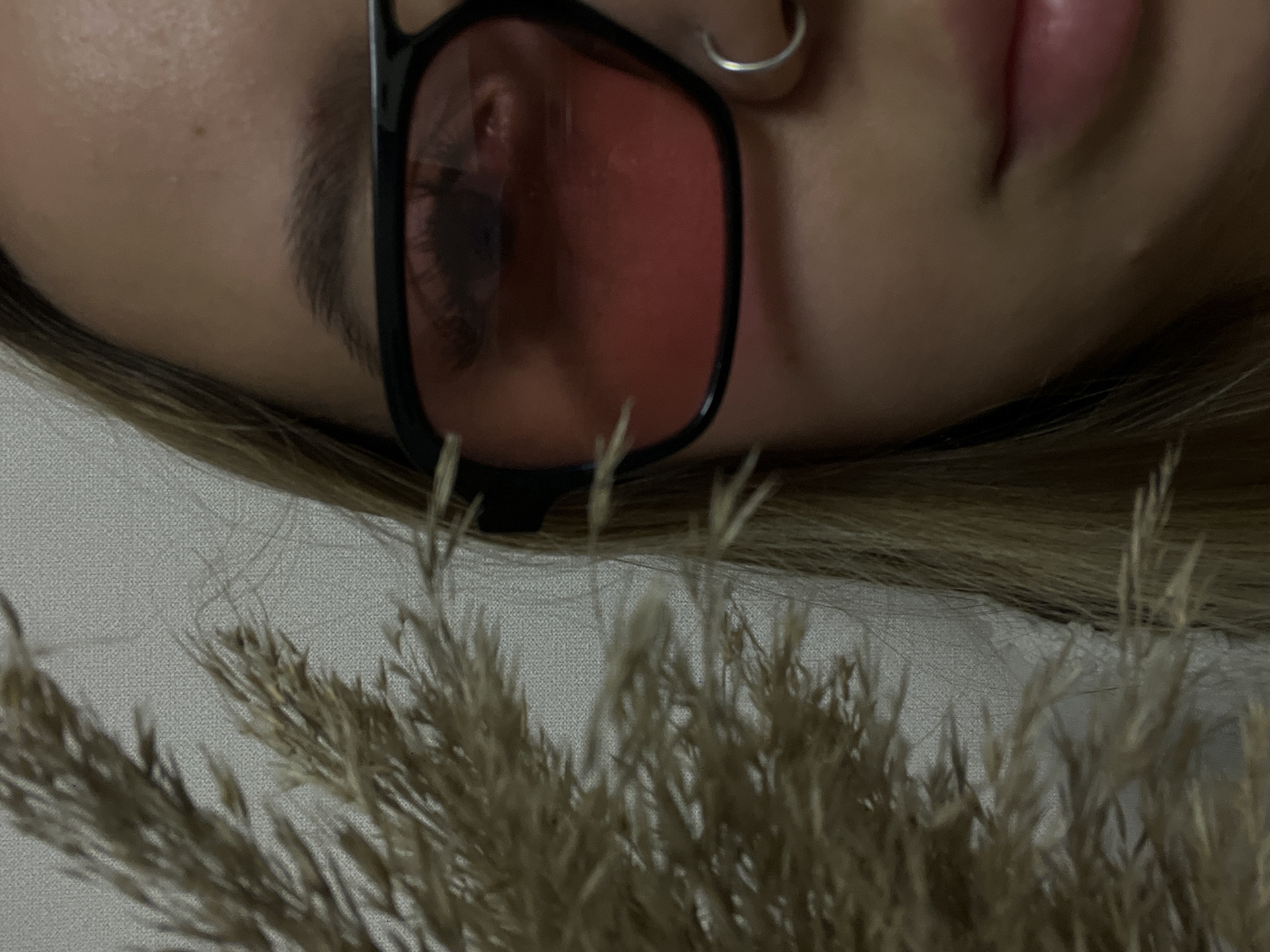 A close-up, rotated shot captures a person's upper face, seen from a slightly overhead perspective. The person, likely female, is lying down, with light-colored hair fanning out around their head. They are wearing black-framed glasses with distinctive reddish-pink tinted lenses, through which a portion of their eye and eyebrow are visible. A simple silver-colored hoop earring adorns the visible earlobe. Below the glasses, part of the nose and softly parted lips are discernable.
In the foreground, a prominent cluster of dried, feathery grass (resembling pampas grass) is out of focus, adding a soft, textural element to the composition. The background is a plain, light-colored, subtly textured surface, possibly a pillow or blanket, indicating an indoor setting. The lighting is soft and even, suggesting an intimate atmosphere. No specific activity is taking place beyond the person appearing to be at rest. There is no visible text.