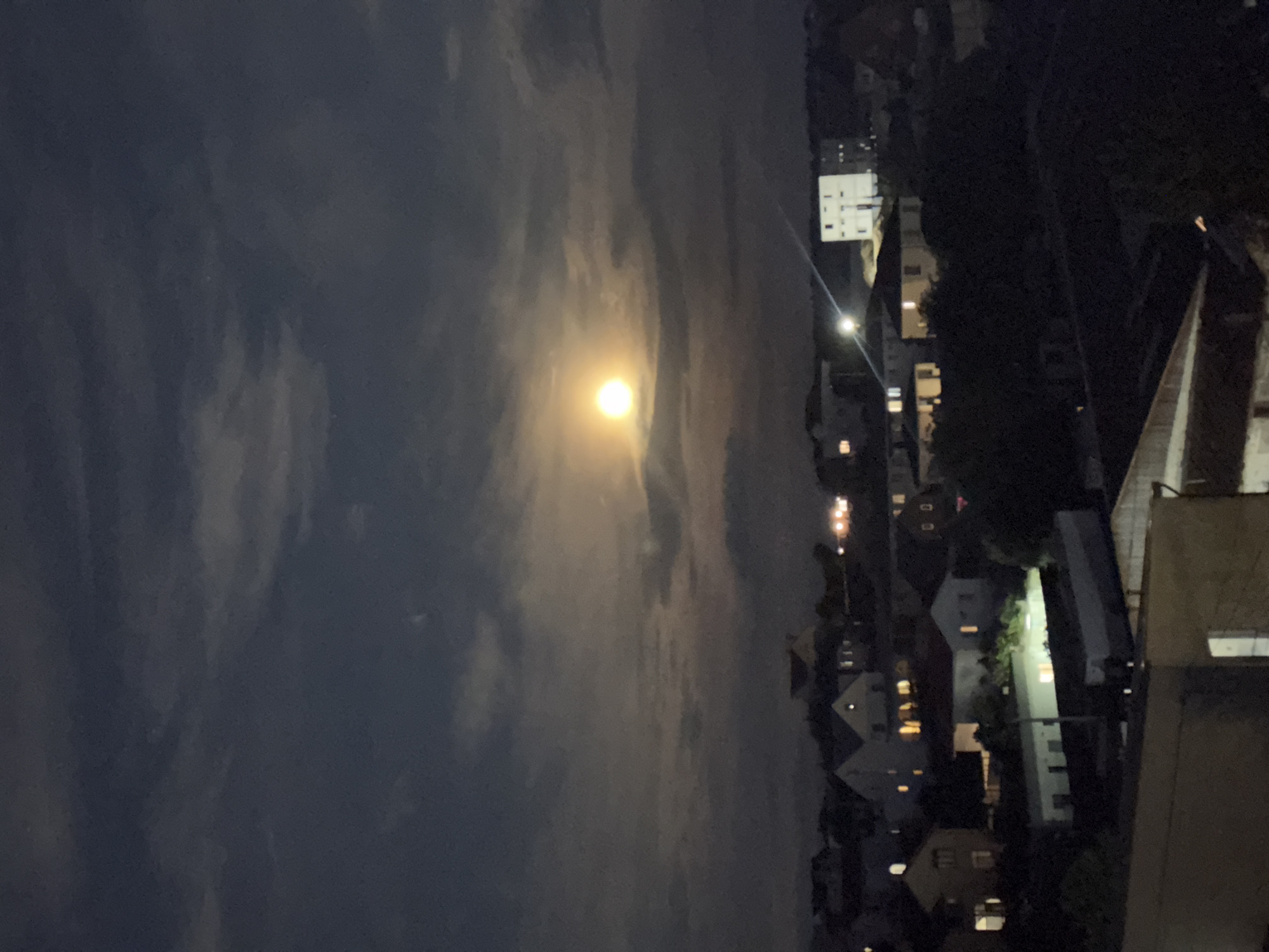 The image shows a nighttime view of a residential area in Balaklavsky District, Ukraine. A bright, full moon is prominently visible in the dark, cloudy sky, illuminating the scene below. Numerous houses, some with lit windows, are scattered across the landscape, indicating an inhabited area. The architecture appears to be typical of a suburban or town setting. Trees and other vegetation are also visible, contributing to the overall landscape. The lighting conditions suggest it is late at night, with the moon being the primary light source. No people or specific actions are discernible in the image.