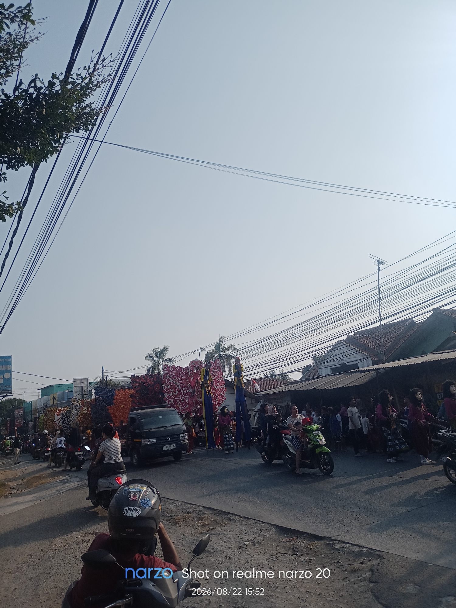 A street scene in Karawang, Indonesia, is depicted under a clear sky, with numerous overhead power lines dominating the upper portion of the frame. The street is lined with buildings and trees. A procession appears to be underway, with several individuals on motorcycles and a black van driving on the road. Large, colorful decorative floats, possibly for a festival or parade, are visible in the middle ground. Some people are walking on the side of the street, and others are on the floats. One float features figures on stilts. The overall impression is a bustling street event.