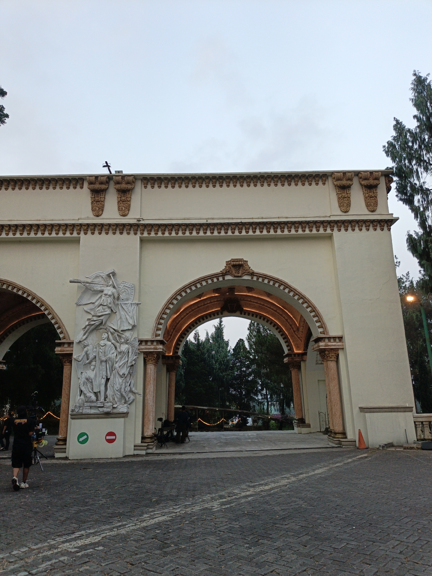 The image shows a wide architectural archway. The arch is cream-colored with gold ornate trim along the top and framing the arch opening. The interior of the arch is lit with warm-toned lights, illuminating the trees visible beyond. On the left side of the archway, there is a white relief sculpture depicting several figures, including winged figures and a central group of three. Below the sculpture are two circular signs, one green with a white arrow pointing left, and one red with a white horizontal bar.
In the foreground, a person wearing a black t-shirt and shorts is walking away from the camera, carrying camera equipment mounted on a tripod. The ground is paved with dark cobblestones. To the right of the archway, a modern street lamp is visible, and further back, trees and some decorative lights can be seen. The sky is overcast and light gray. The scene appears to be an outdoor location, possibly a public area or attraction in Cibogo, Indonesia.