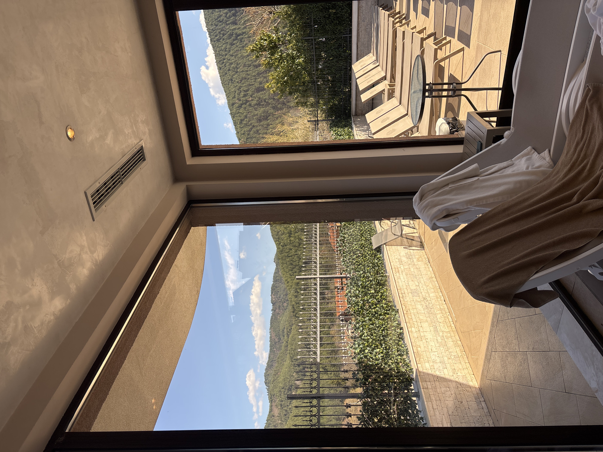 A low-angle interior shot captures a view through two rectangular windows. The wall is a neutral beige, and a rectangular vent is visible in the upper-left. The upper window offers a view of a forested mountain range against a blue sky, with outdoor furniture and a circular table visible in the foreground. The lower window shows a similar mountain view, but with a decorative black fence in the foreground and a brick wall. Part of a white chair with towels is in the right foreground. The location is Unknown, Unknown.