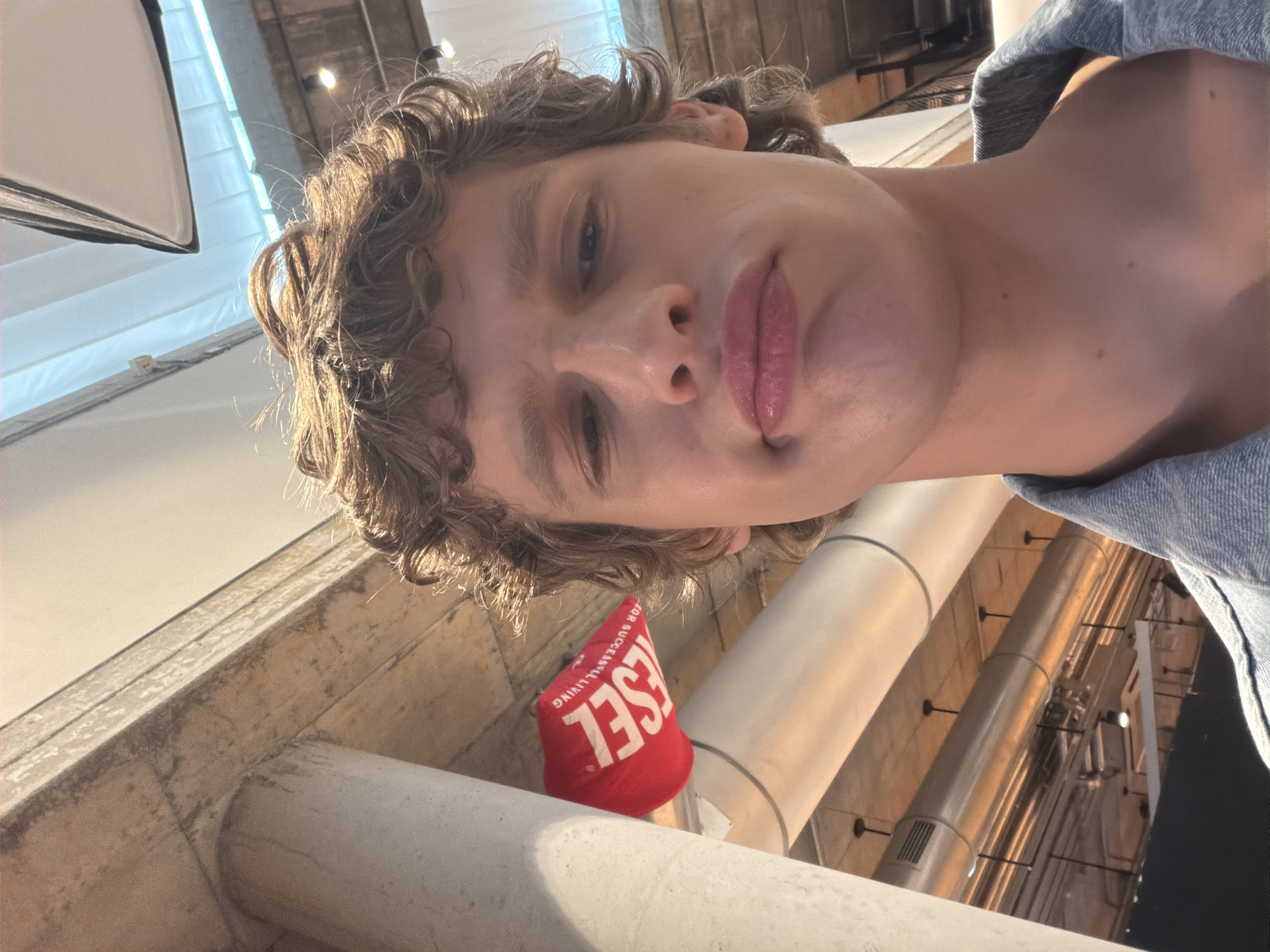 The image is a low-angle selfie of a person with light skin, curly hair, and pursed lips, positioned indoors. The background includes architectural elements like support pillars and overhead pipes. A small red object with white lettering, potentially a promotional item or advertisement, is placed prominently near the bottom of the frame. The lighting is bright, possibly from studio lights or large windows. The person is wearing a denim shirt. The location appears to be an industrial-style space. It is not possible to know the city, Unknown, Unknown from the available information.