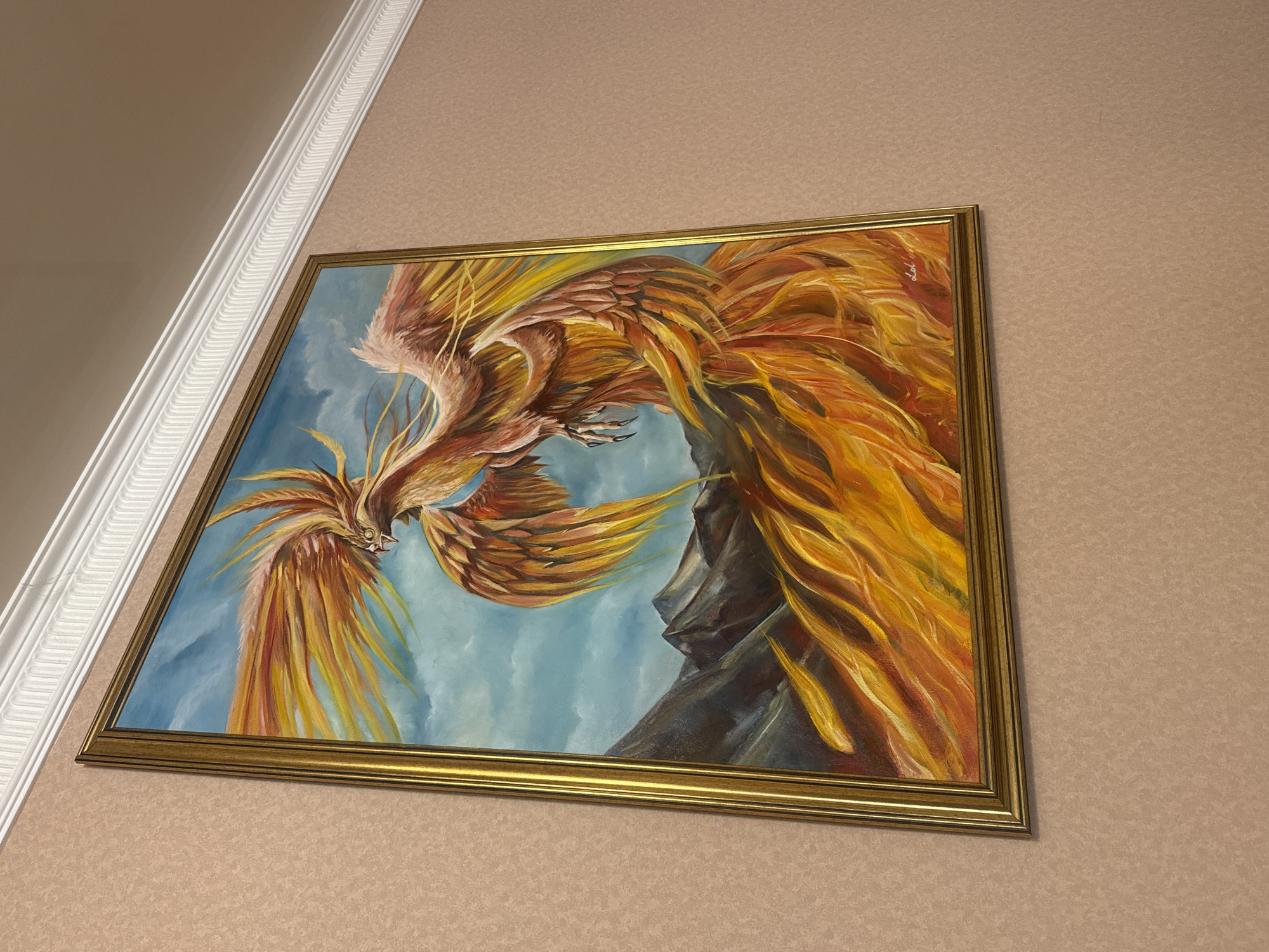 A framed painting of a phoenix hangs on a wall, likely indoors. The painting depicts a phoenix with fiery, primarily orange and yellow, plumage against a cloudy blue sky. There's a gray rock formation visible in the background of the painting. The frame is gold. The wall has a textured, light brown/beige wallpaper. A white decorative molding runs along the top of the wall, near the ceiling. The photo was likely taken in Yerevan, Armenia.