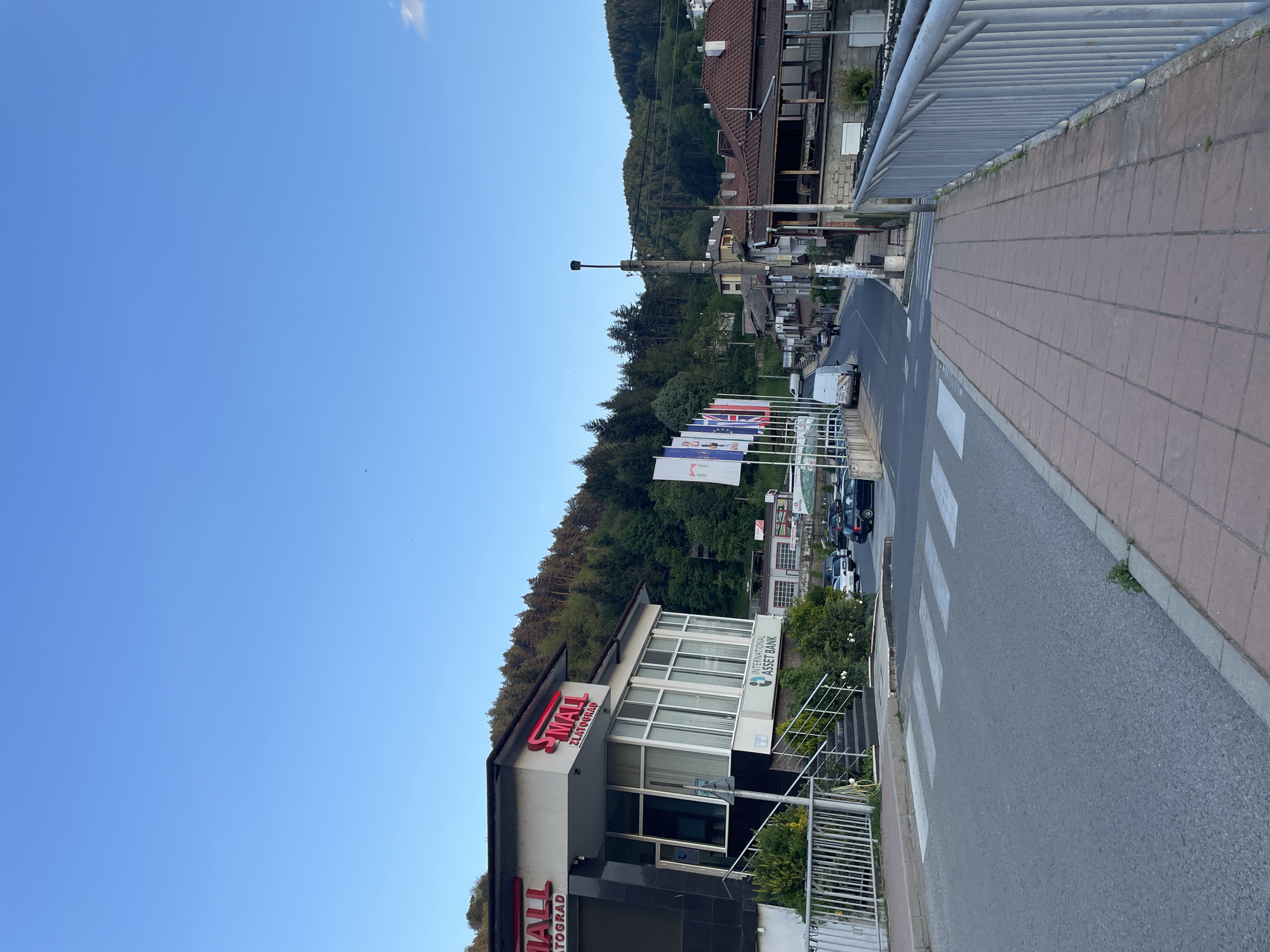 The image depicts a street scene in Zlatograd, Bulgaria. The focus is on the "Small Zlatograd" building, an International Asset Bank branch, located near a crosswalk. Behind the building, numerous flags are visible, lining the street. Cars are parked along the roadside. The background features a forested hillside and residential buildings. The sky is clear blue. The image is taken from an elevated position, likely a bridge or overpass.