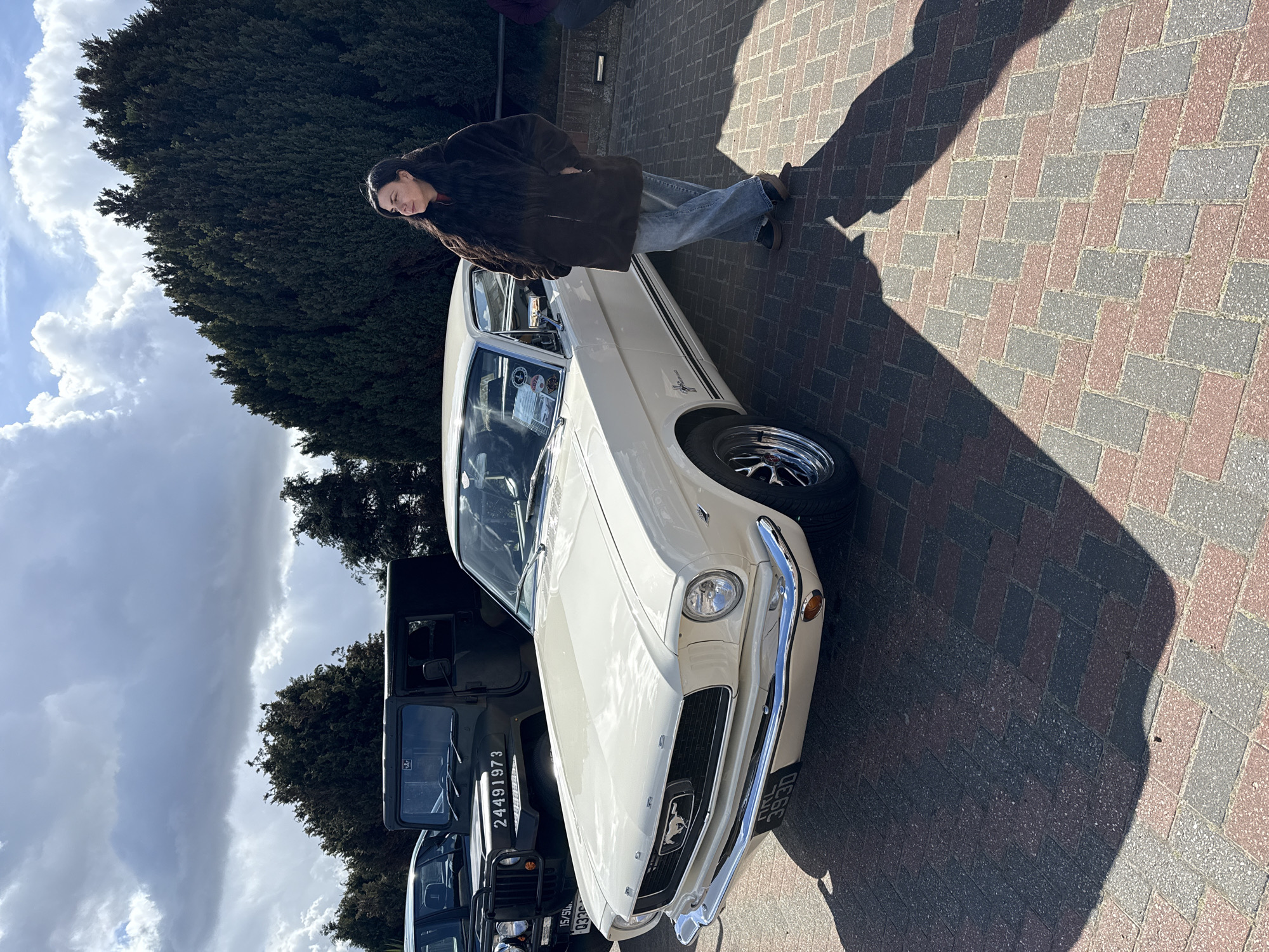A medium shot shows a person standing beside a cream-colored Ford Mustang on a brick-paved area. The individual, dressed in a brown jacket and jeans, is leaning casually against the car. The Mustang is well-maintained and has a classic design, indicative of the mid-1960s era. The license plate, "DRL 393D," is visible on the front. Behind the Mustang, partially obscured by trees, are two military vehicles, one with the number "24491973" on the windshield. The environment includes overcast skies, suggesting a cloudy day. The location is not explicitly stated as Great Yarmouth, but the presence of UK license plates and military vehicles suggests it is somewhere in the United Kingdom.