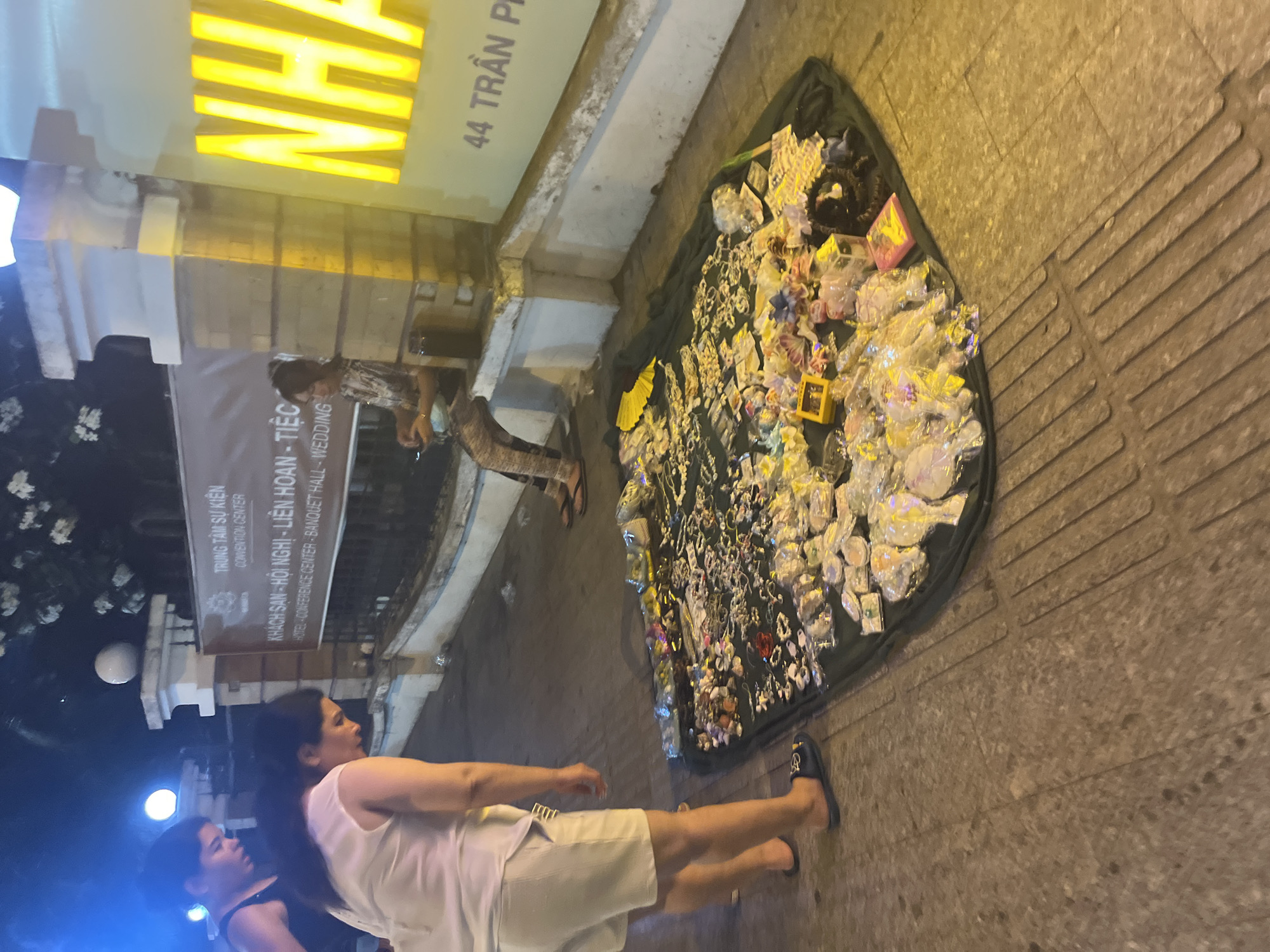 In Nha Trang, Vietnam, two individuals are observed on a sidewalk at night. One person, wearing a black tank top and dark pants, is standing and looking down. The other person, dressed in a white tank top and white shorts, is also looking downwards towards a display of merchandise spread out on a dark cloth on the ground. The merchandise includes an assortment of small items, possibly jewelry and accessories, some of which are packaged in clear plastic. A third individual, wearing a patterned top and pants, is seated on a low wall behind the display, looking down at their hands, with a mask resting on their lap. Behind them, a building facade features illuminated signage with Vietnamese text and the number "44 Trần Phú". The environment suggests a street vendor setup with goods laid out for sale.
