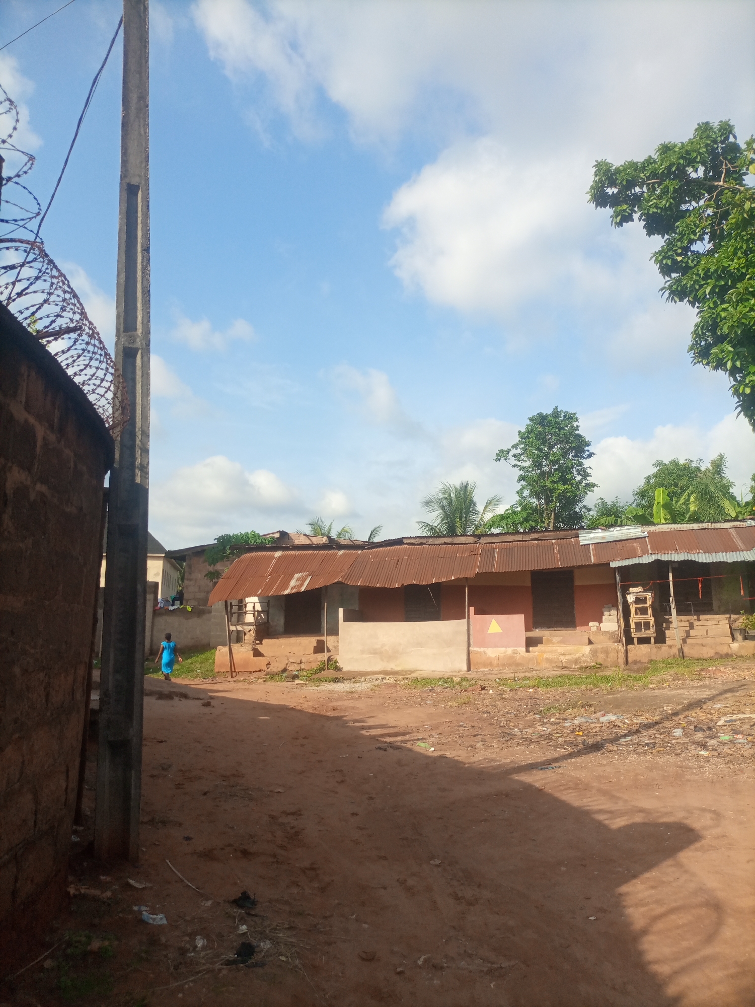 The image depicts a street scene in Nkpor, Nigeria, on a bright, partly cloudy day. The sun casts long shadows across the dusty, unpaved road, suggesting it is either early morning or late afternoon.
In the foreground, a tall, grey concrete pole stands to the left, partially obscured by barbed wire fencing running along a brick wall. The wall, constructed from rough, reddish-brown bricks, dominates the left side of the frame.
Further down the road, a person wearing a bright blue dress walks away from the viewer towards a cluster of buildings. These buildings appear to be modest, with corrugated metal roofs showing signs of age and rust. The walls of the structures are a pale, earthy color. One building has a small, yellow triangular marking on its exterior. In front of the buildings, there are steps leading up to an entrance. Some wooden structures, possibly shelving or supports, are visible. Greenery, including trees and grass, is present in patches around the buildings.
The overall scene conveys a sense of everyday life in a semi-urban or rural environment. The presence of debris and litter on the road indicates potential waste management challenges. The clear sky and bright sunlight suggest fair weather. No visible text is present in the image.