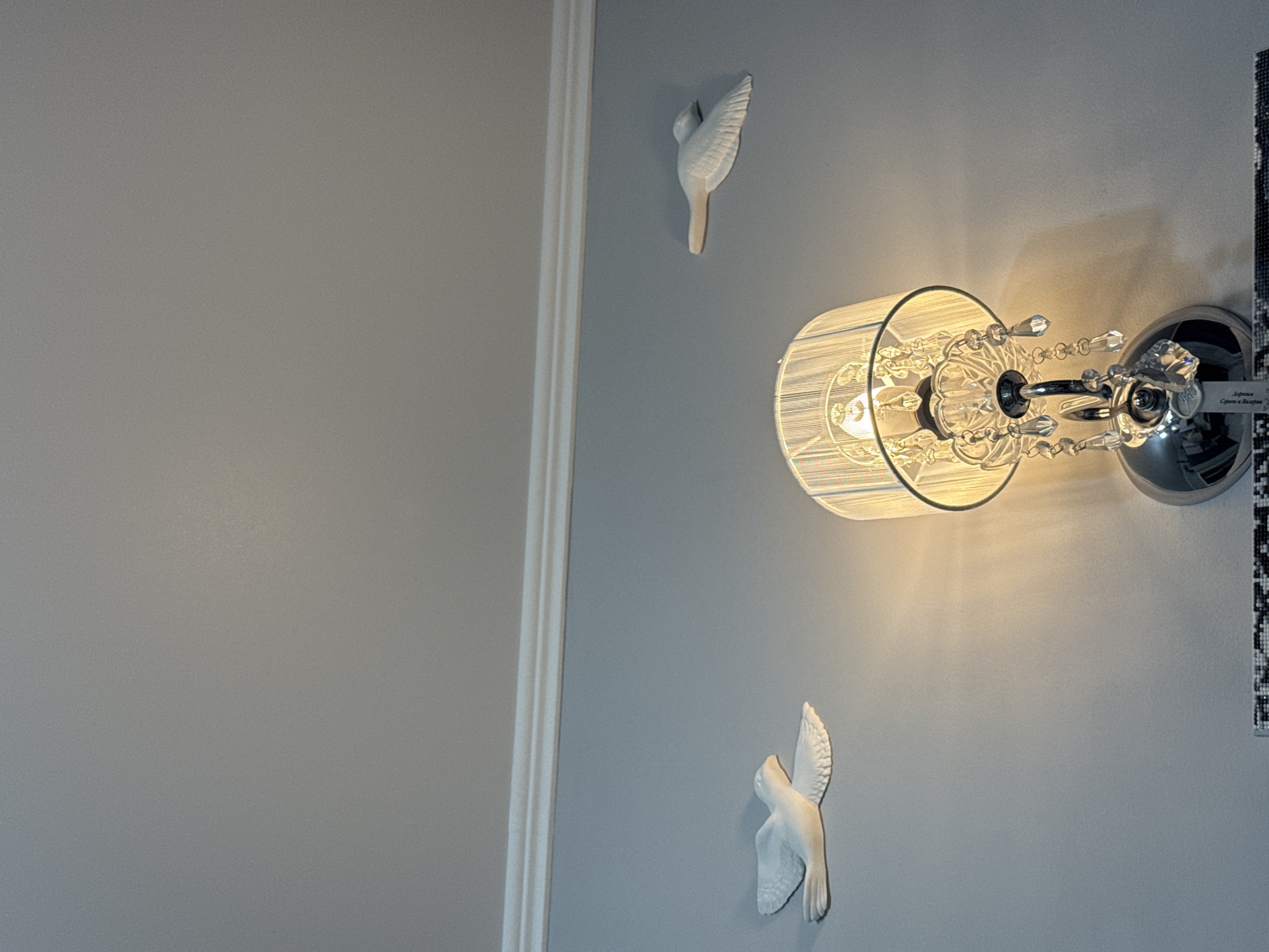 The image captures a close-up, angled view of a light blue-gray wall adorned with decorative elements. The central subject is a wall-mounted light fixture (sconce) with a cylindrical, translucent white shade that is illuminated, casting a warm, soft glow on the wall. The fixture features clear, multifaceted crystal embellishments hanging from its chrome-finished base.
Flanking the light fixture are two white, sculpted bird decorations, resembling doves, affixed to the wall. One bird is positioned above the light, and the other is below, both appearing to be in flight. To the left, a white vertical strip of architectural molding or trim runs from top to bottom, separating the light blue-gray wall from an adjacent, slightly darker gray wall area.
On the far right edge of the image, partially visible, is what appears to be a framed piece of artwork or a detailed decorative panel. Attached near the light fixture, on a small label or plaque, there is visible text in Cyrillic script: "Афины" (Athens) and "Серебро и Балерин...", with the last word truncated. Below this, smaller, less legible text is also present. The scene depicts an interior setting, likely a room, with the artificial light suggesting evening or an indoor space without much natural light.