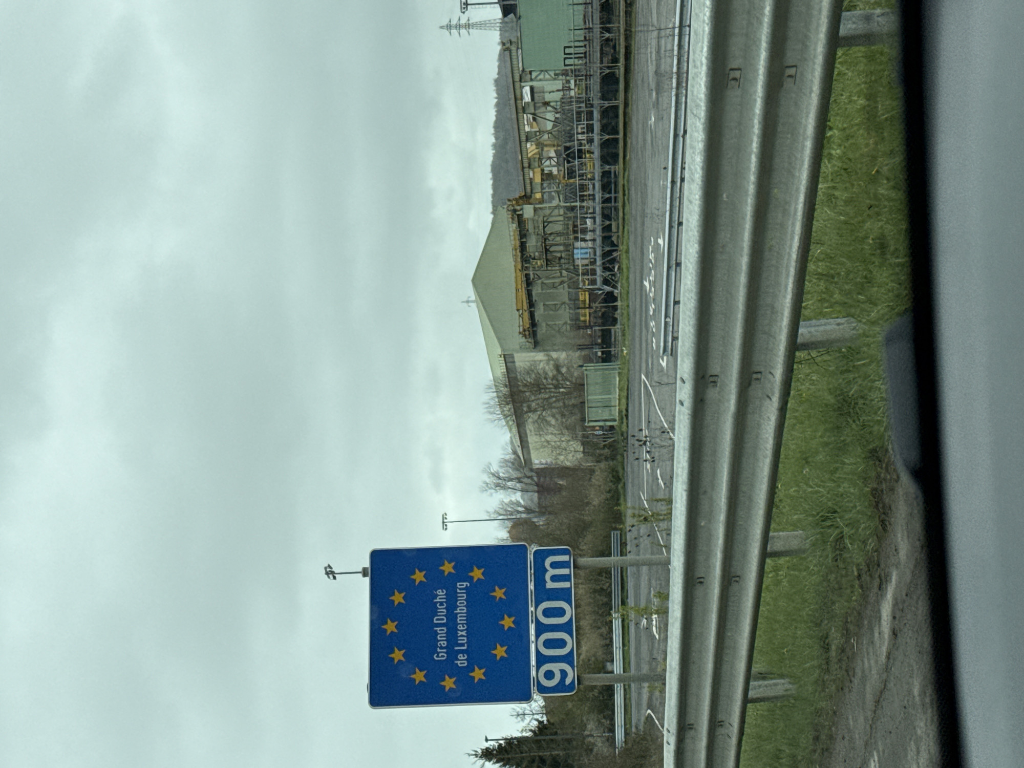 The image shows a road sign indicating the border to Luxembourg (Grand Duche de Luxembourg) is 900 meters away. The sign is blue with yellow stars, typical of EU country border signage. A metal guardrail is visible on the right side of the road. Behind the sign, there's a large industrial complex or factory, with tanks and scaffolding. The sky is overcast. The location is likely near Aubange, Belgium, given its proximity to the Luxembourg border.