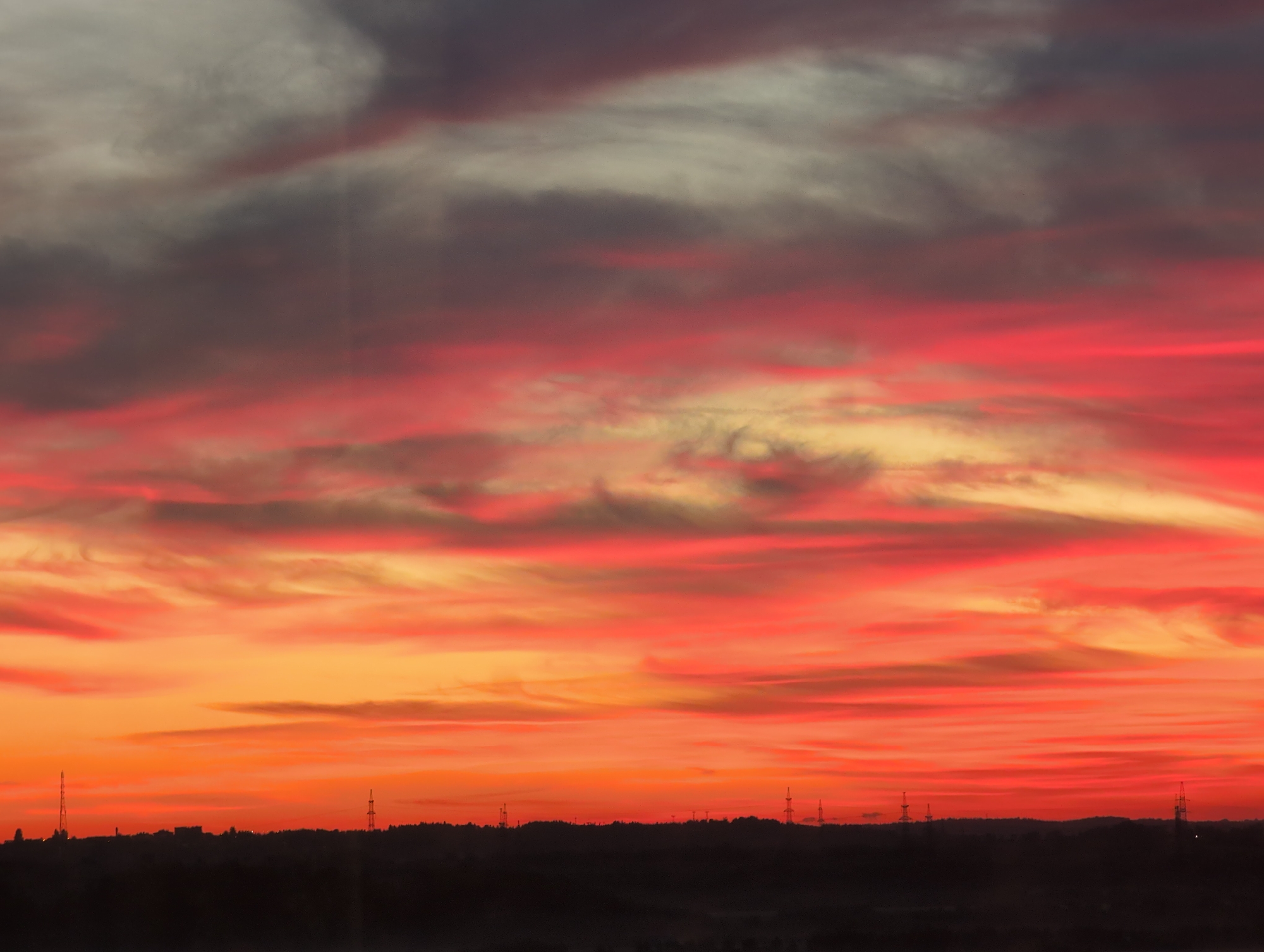 The image captures a dramatic sunset over the city of Bugry, Russia. The sky is ablaze with vibrant hues of red, orange, and yellow, painted with wispy clouds that stretch across the frame. A dark, silhouetted horizon line marks the edge of the city, with buildings and communication towers faintly visible against the colorful sky. The overall impression is one of natural beauty and the serene transition from day to night.