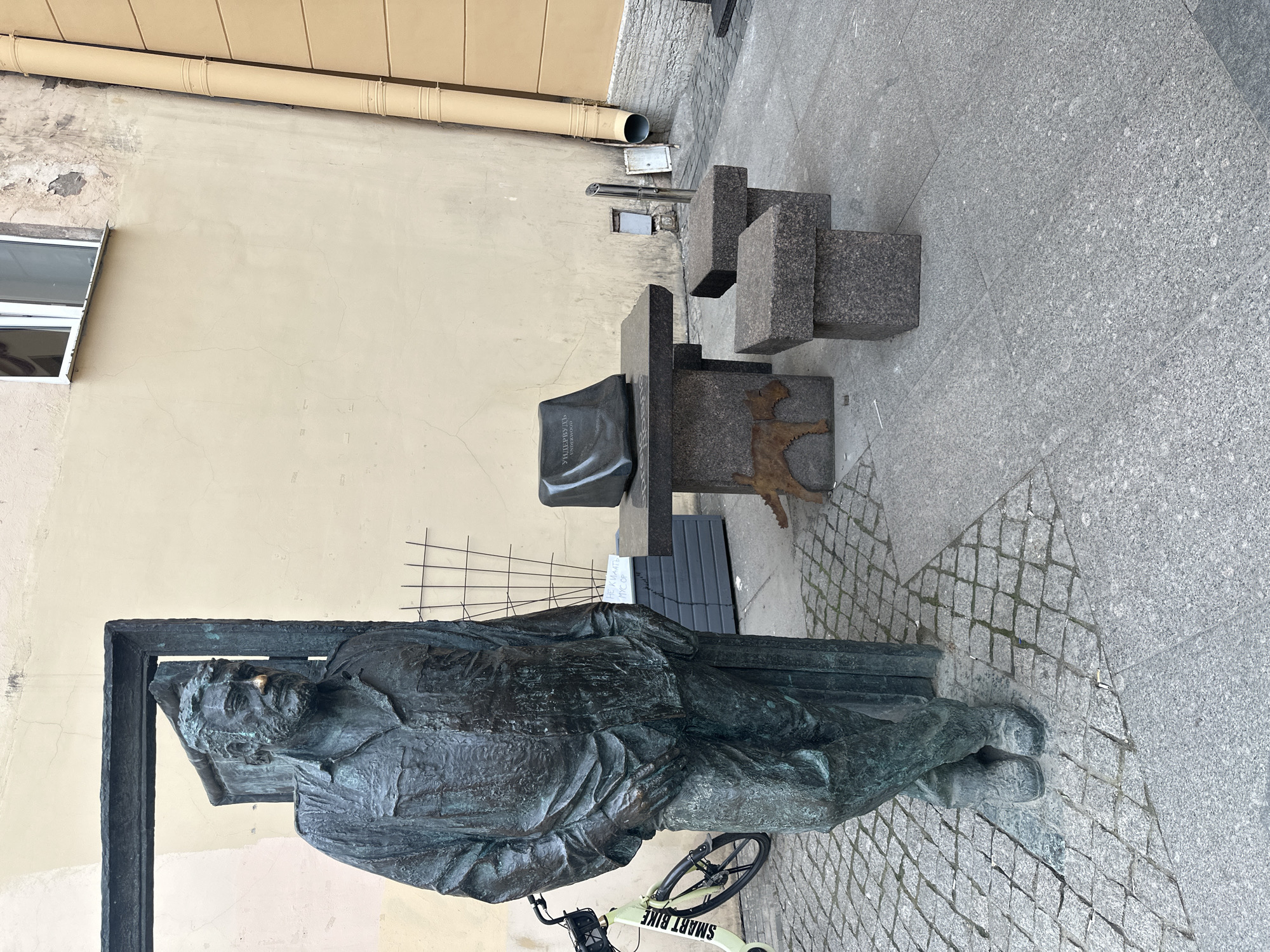 The image shows the monument to Sergei Dovlatov, located on Rubinstein Street in Saint Petersburg, Russia. The bronze statue depicts the writer reclining on a bench, seemingly lost in thought. Next to the bench is a stylized monument made of granite blocks, including a depiction of a dachshund. A bicycle is parked near the statue's feet. The background consists of a building wall and a paved sidewalk.