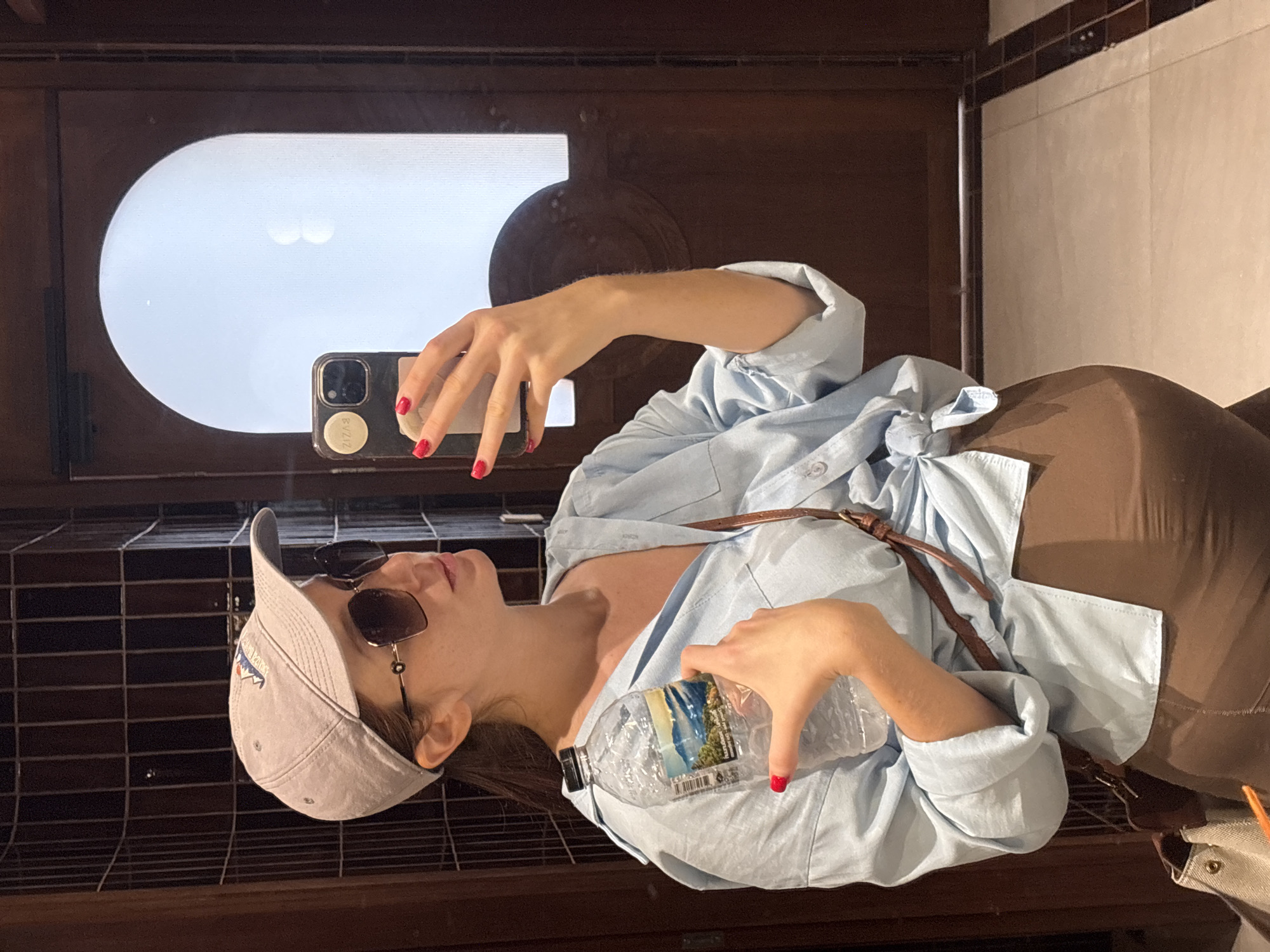 A woman in a light blue shirt, brown pants, and a light-colored baseball cap is taking a selfie in a bathroom in Choeng Thale, Thailand. She is holding a water bottle in one hand and a smartphone in the other, with red nail polish visible on her fingers. She is wearing sunglasses. The bathroom features brown wooden cabinets with tiled backsplashes. A window or mirror with a rounded rectangular shape is visible behind her phone. The background includes beige tiles.