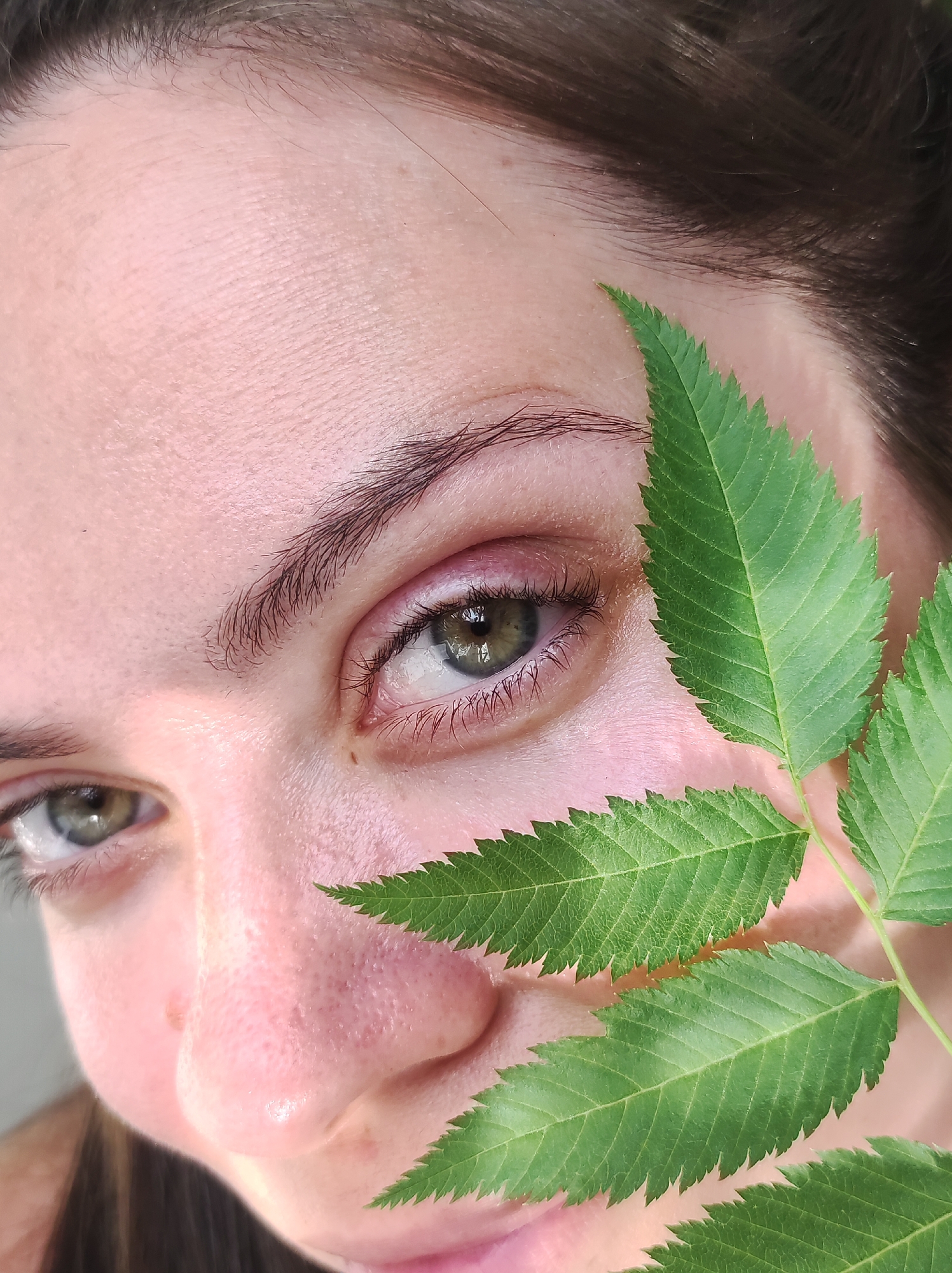 A close-up image shows a portion of a person's face with a green leaf covering part of it. The person has fair skin, brown hair, and green eyes. The left eye is fully visible, while the right eye is partially obscured by the leaf. The leaf is green with serrated edges. The setting and any location in Chaikovsky, Russia is not evident in the image.