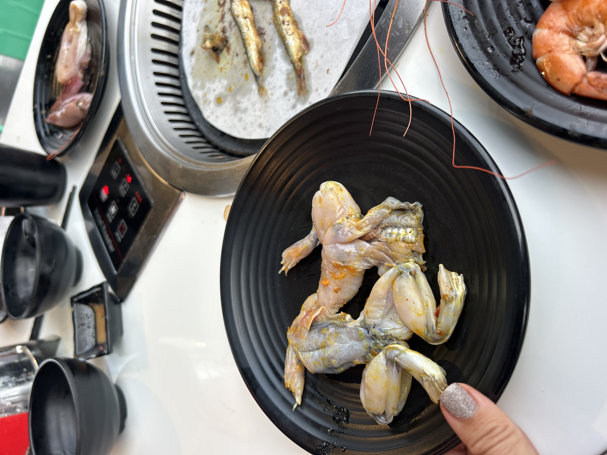 This close-up, high-angle image captures a vibrant dining scene in Nha Trang, Vietnam, centered around an electric grill. In the foreground, a black ceramic plate holds several raw frog legs, lightly seasoned with spices or marinade, indicating preparation for grilling. A hand with glittery silver nail polish is visible in the lower right corner, seemingly holding or picking up one of the frog legs.
To the upper right, another black plate contains several shrimp, which appear either raw and seasoned or lightly cooked. An electric grill occupies the middle-left of the frame, featuring small fish, possibly anchovies, cooking on a piece of parchment paper placed over its grates. The grill's control panel, with red indicator lights and barely discernible text for settings like "ON/OFF" or temperature control, is visible on its side. Fine reddish-orange wires are loosely draped near the shrimp plate. In the far upper left, a small black bowl contains pieces of raw meat, likely for grilling, and further left, several empty black ceramic cups or bowls are arranged on the white table.
The scene depicts an interactive dining experience, characteristic of Asian barbecue or hot pot meals, where diners cook their food fresh at the table. The array of ingredients, including frog legs, shrimp, and small fish, points to a diverse culinary offering typical of the region. The setting, characterized by individual cooking apparatus and multiple plates of ingredients, suggests a restaurant or communal dining environment in Nha Trang, Vietnam.