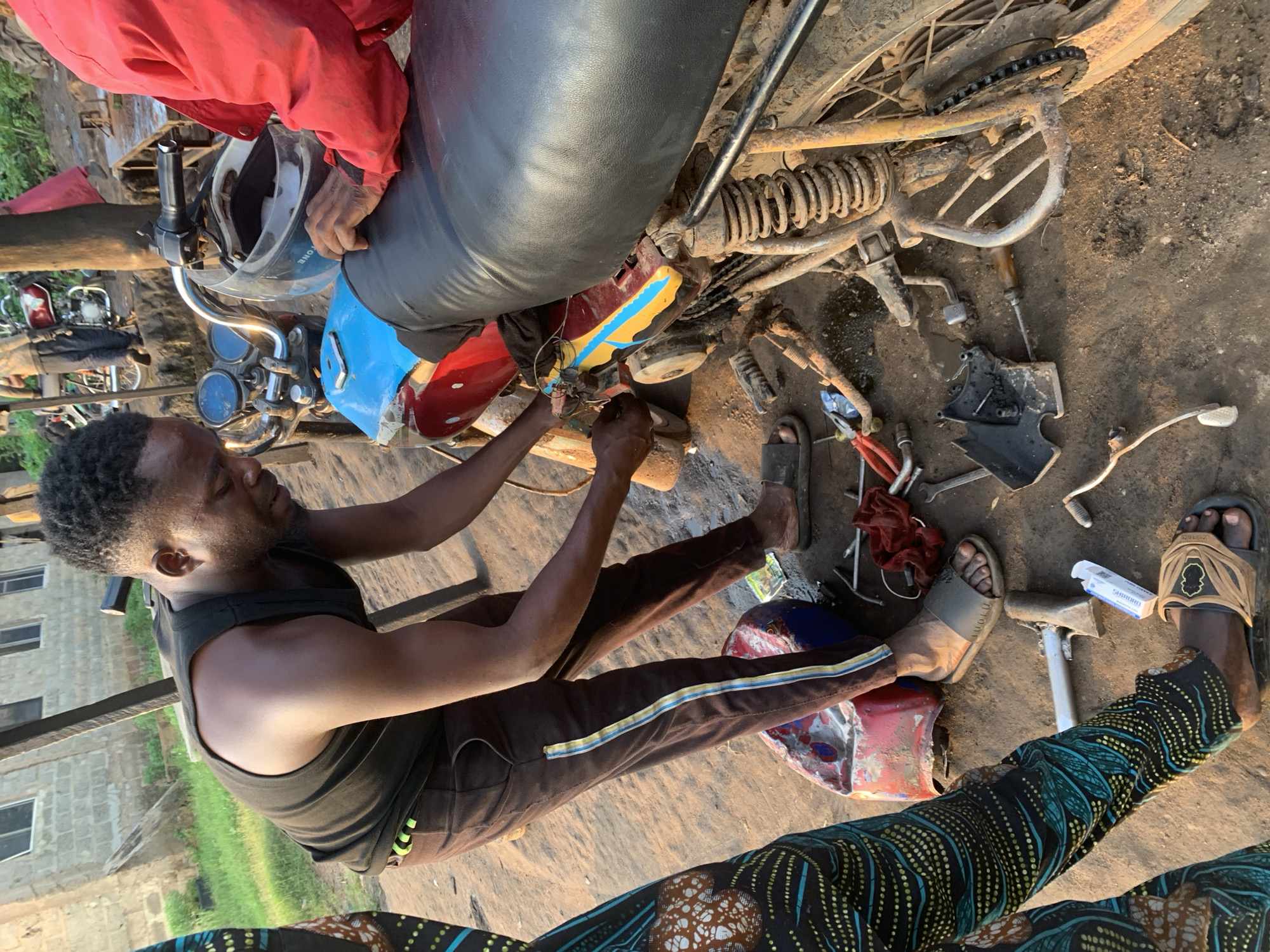 In Shagamu, Nigeria, a Black man wearing a black tank top and brown pants with colored stripes is working on a red and blue motorcycle. He is bending over, focused on the engine area of the motorcycle, with his hands on the mechanism. Tools are scattered on the dirt ground around the motorcycle, including pliers, a screwdriver, and a red cloth. A red and blue plastic container is also visible on the ground near his feet. Another person, partially visible on the left side of the frame, wears a colorful patterned garment. In the background, a brick building with windows and wooden posts suggest an outdoor workshop setting.