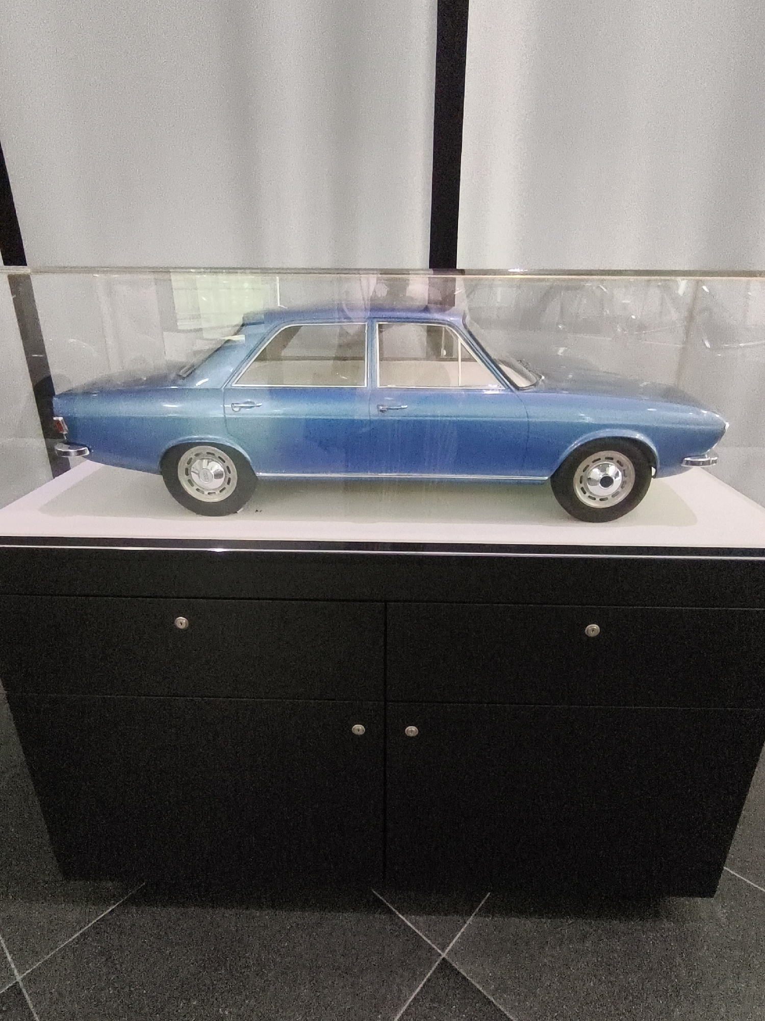An illuminated scale model of a blue sedan car is displayed inside a clear protective case. The car model is positioned on a white surface, which sits atop a black cabinet with two doors, each featuring a silver handle. The cabinet is located on a dark grey, textured floor. The background consists of a light grey, textured wall with a vertical black element on the left side. The scene appears to be in a well-lit exhibition or showroom. This display is located at the Audi Forum Ingolstadt in Ingolstadt, Germany, a facility dedicated to the Audi brand, showcasing its history, design, and technology.