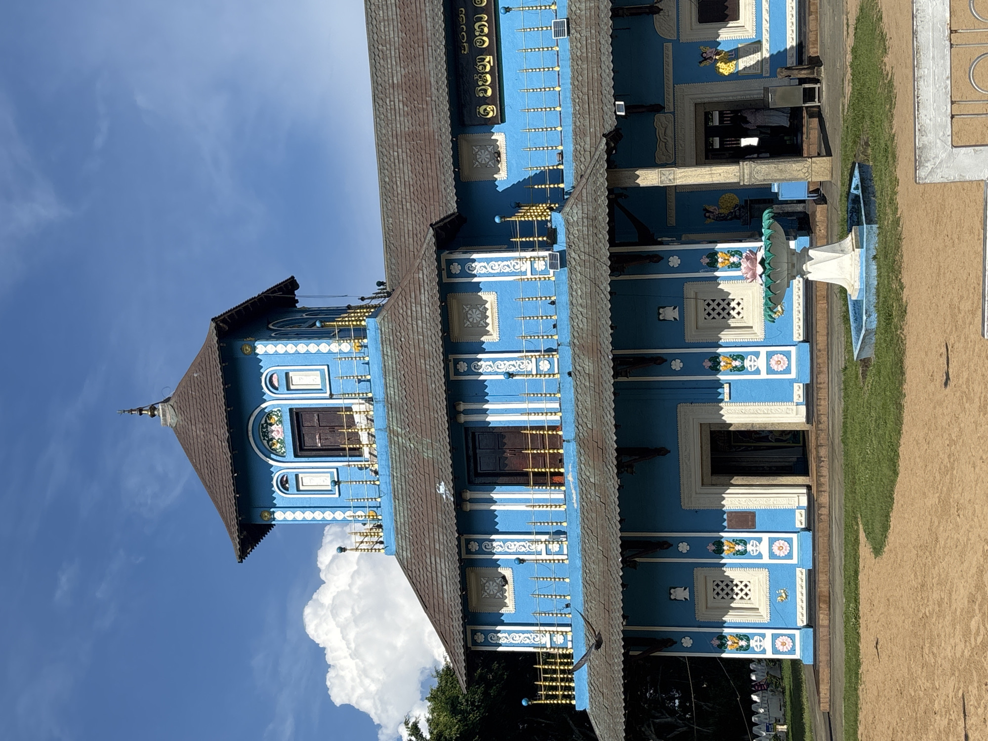 The image shows the facade of the Sri Vishnu Maha Devalaya in Dondra, Sri Lanka. It is a blue building with a multi-tiered roof. The architecture is ornamented with white trim, floral motifs, and decorative lattice windows. A white fountain sits in front of the building. The sky is blue with puffy white clouds behind the building. The ground is covered with grass and sand.