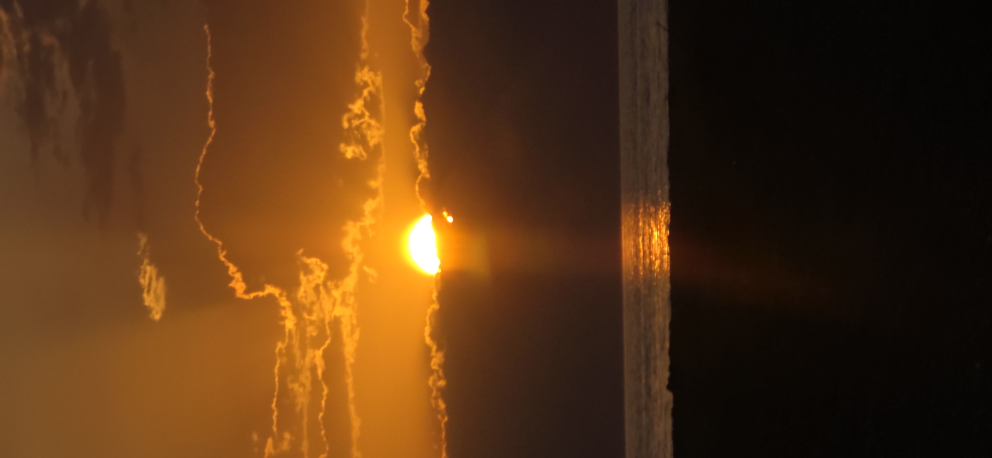 This is a vertical, medium-shot photograph of a sunset over a body of water, possibly a sea or large lake, viewed from a sandy shore. The sun, a bright, glowing orb, is partially obscured by dark, horizontal clouds, with rays of light emanating downwards. The sky above is a gradient of warm oranges and yellows, with smaller, illuminated clouds scattered throughout. The water reflects the vibrant colors of the sunset, creating a shimmering pathway of light. The foreground consists of a dark, silhouetted expanse, likely the shore or beach, with some indistinct texture. There are no people or specific structures visible in the image. The overall scene evokes a peaceful and natural ambiance.