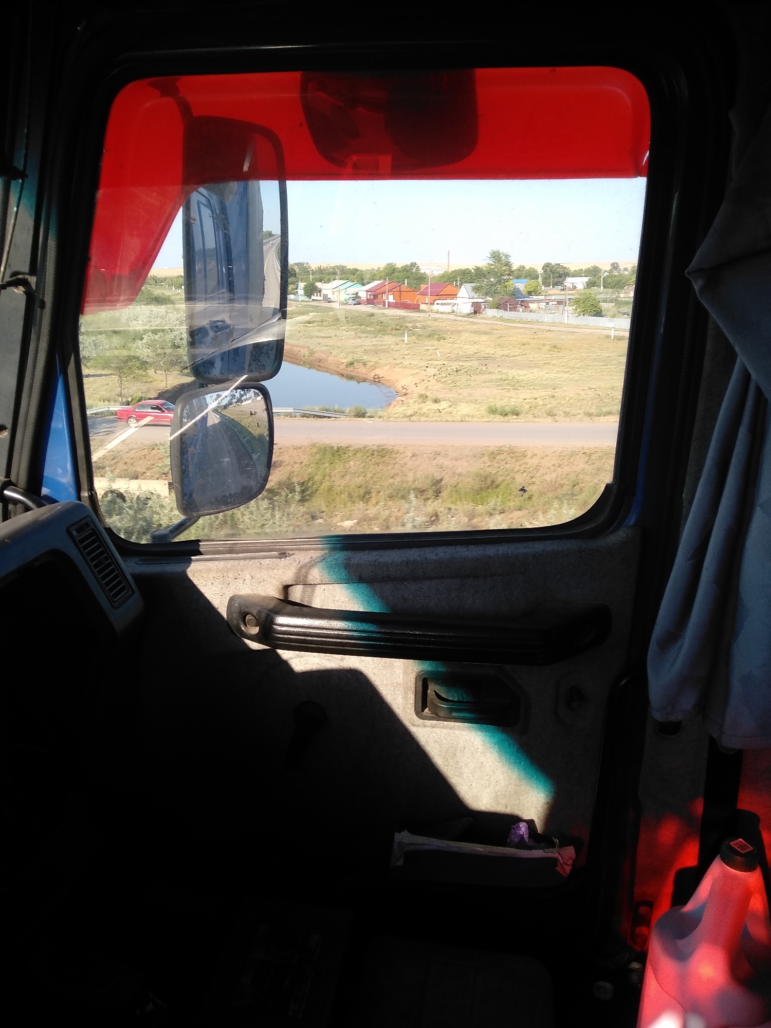 The image is taken from inside the cab of a truck, looking out the driver's side window. The window features a red sun visor. Outside the window, a rural landscape is visible, with a body of water, dry grass, a road, and several houses with red roofs. A red car is parked on the side of the road. The interior of the truck cab is visible in the foreground, showing the door panel with a handle and a vent. The lighting suggests it is daytime. The image captures a view of the outskirts of the village of Batpakty, Kazakhstan, from a truck.