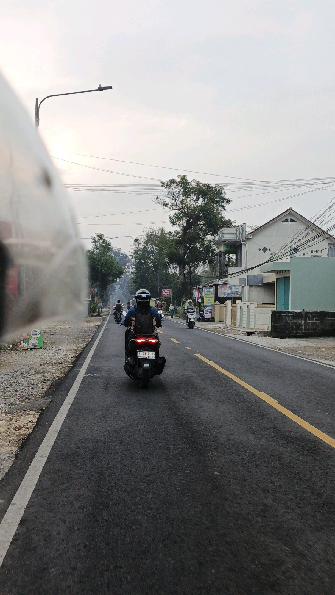 The image captures a street scene in Sukaresik, Indonesia, looking forward from a motorcycle. The road is paved with asphalt and features a yellow dividing line. On the right side of the road, several buildings are visible, including houses and shops. Lush green trees line the street, and power lines crisscross overhead. In the foreground, a person is riding a black motorcycle, wearing a helmet and a backpack. Other motorcyclists are also present on the road, moving in the same direction. The sky is overcast, suggesting either early morning or late afternoon. A street lamp is also visible. The overall impression is that of a typical day in a suburban or semi-urban area of Sukaresik.