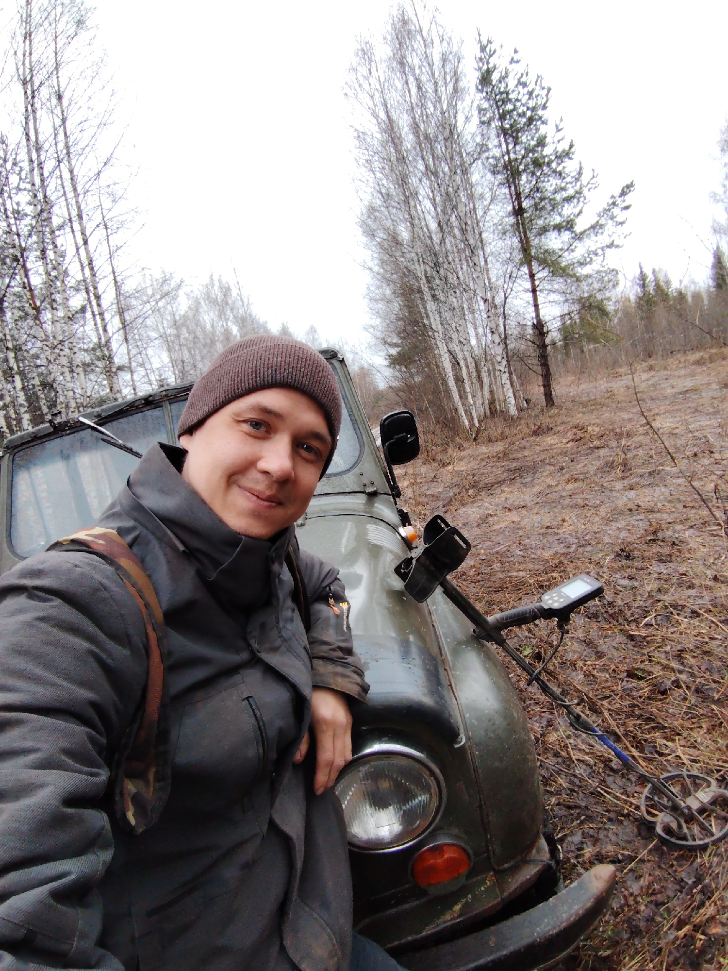 A medium shot captures a man in a brown beanie and dark jacket, standing next to a green military-style vehicle in what appears to be a rural, wooded area. The man has a slight smile and is leaning on the front of the vehicle near the driver's side headlight. A metal detector rests on the vehicle’s hood, with its circular search coil on the ground. Bare trees populate the background, suggesting a late autumn or early spring season. The ground is covered with dried leaves and damp vegetation. The city of Markovskii, Russia is not readily evident in the image.